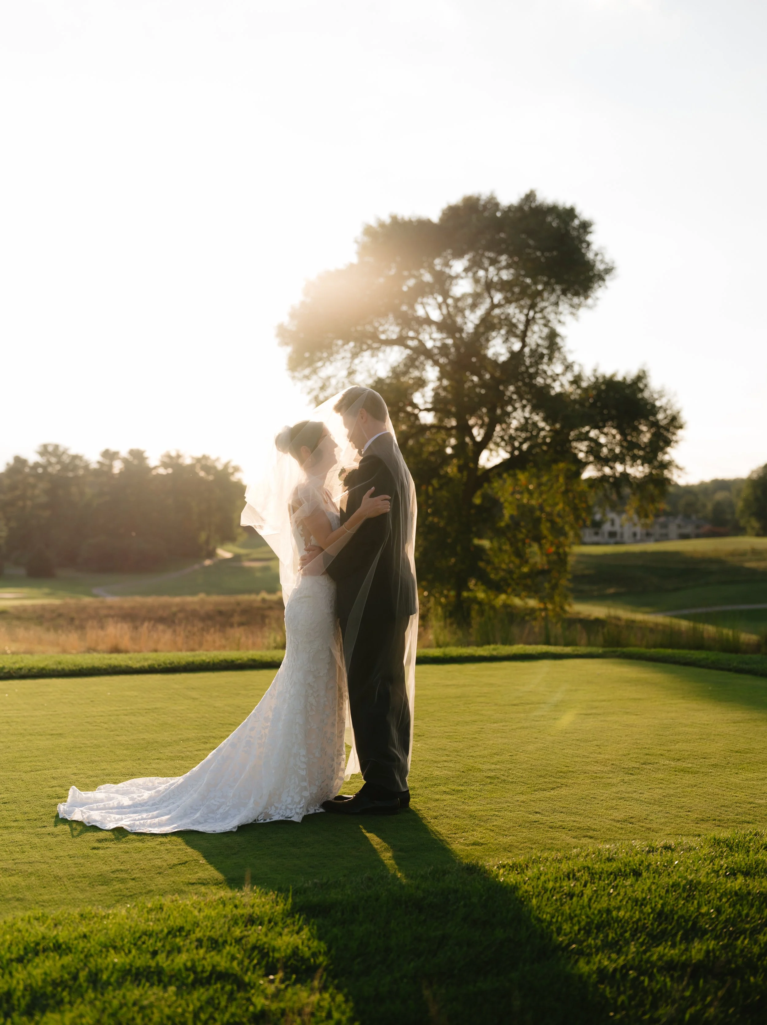 A bride and groom stand close together on a green lawn with the sun setting behind them, sharing an intimate moment.
