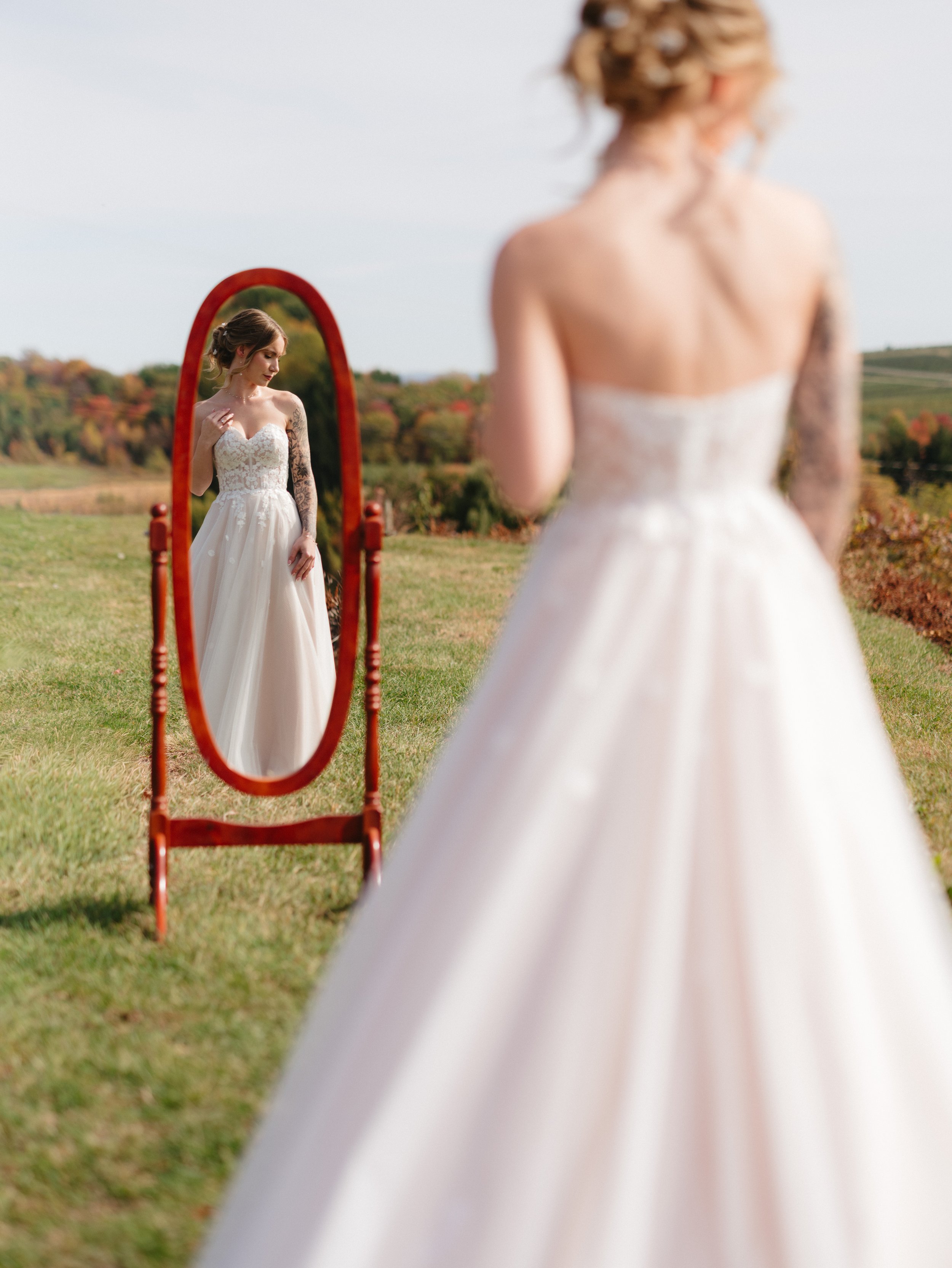 A woman in a wedding dress looks at herself in a standing mirror outdoors on a grassy field, with a scenic landscape in the background.