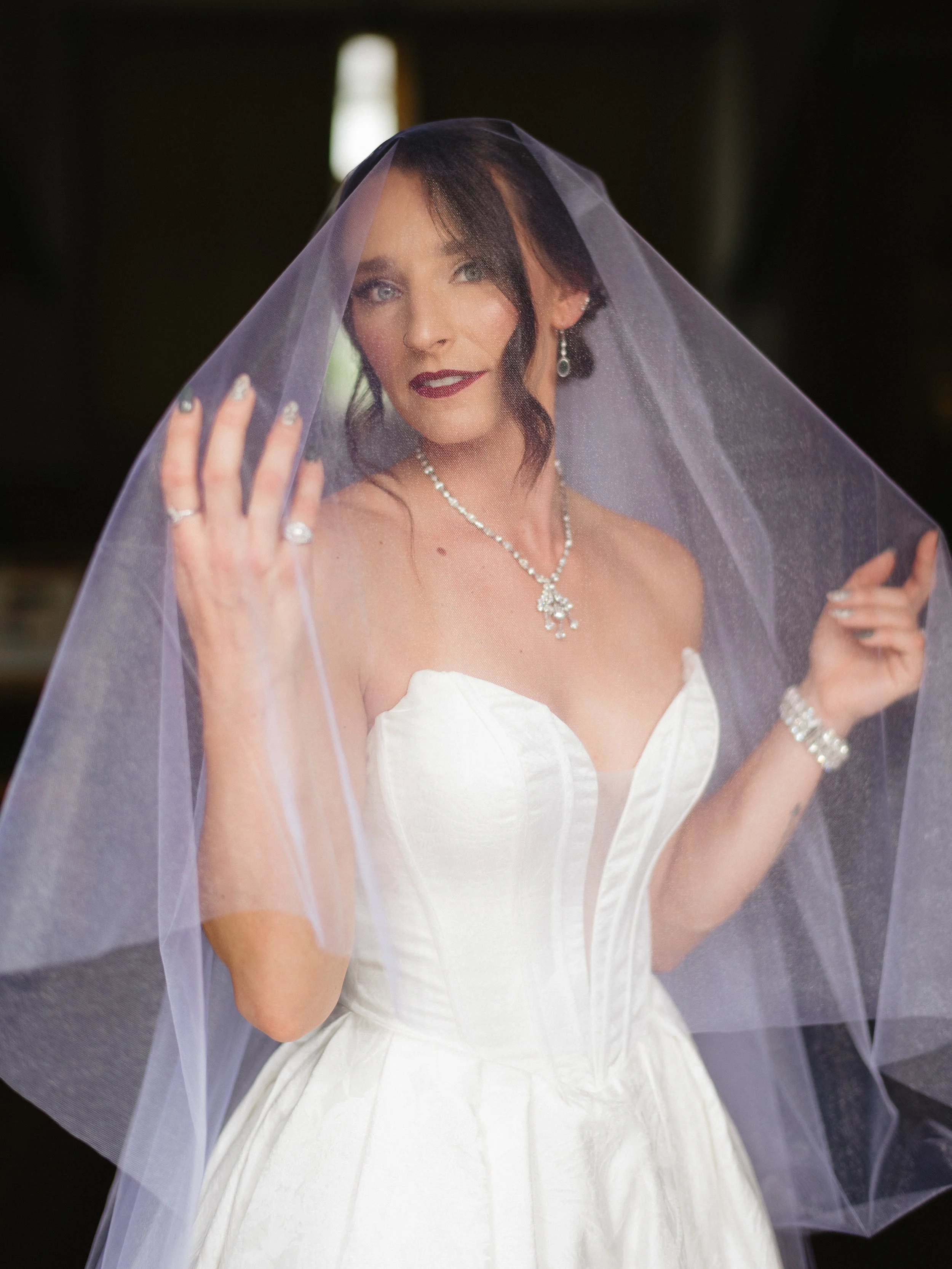 Woman in a white wedding dress with a veil, wearing jewelry, standing indoors with soft lighting. editorial vibes vintage vibes