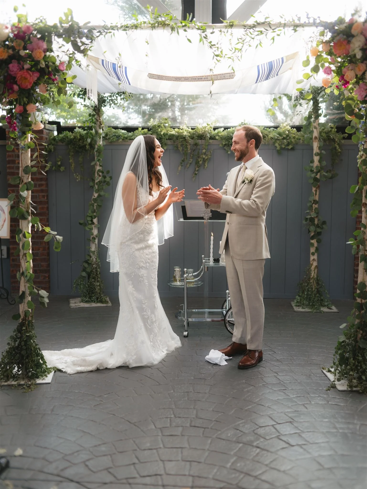 A bride and groom clapping under a chuppah during a wedding ceremony. The groom wears a light-colored suit, and the bride wears a white lace gown and veil. The chuppah is decorated with greenery and flowers.