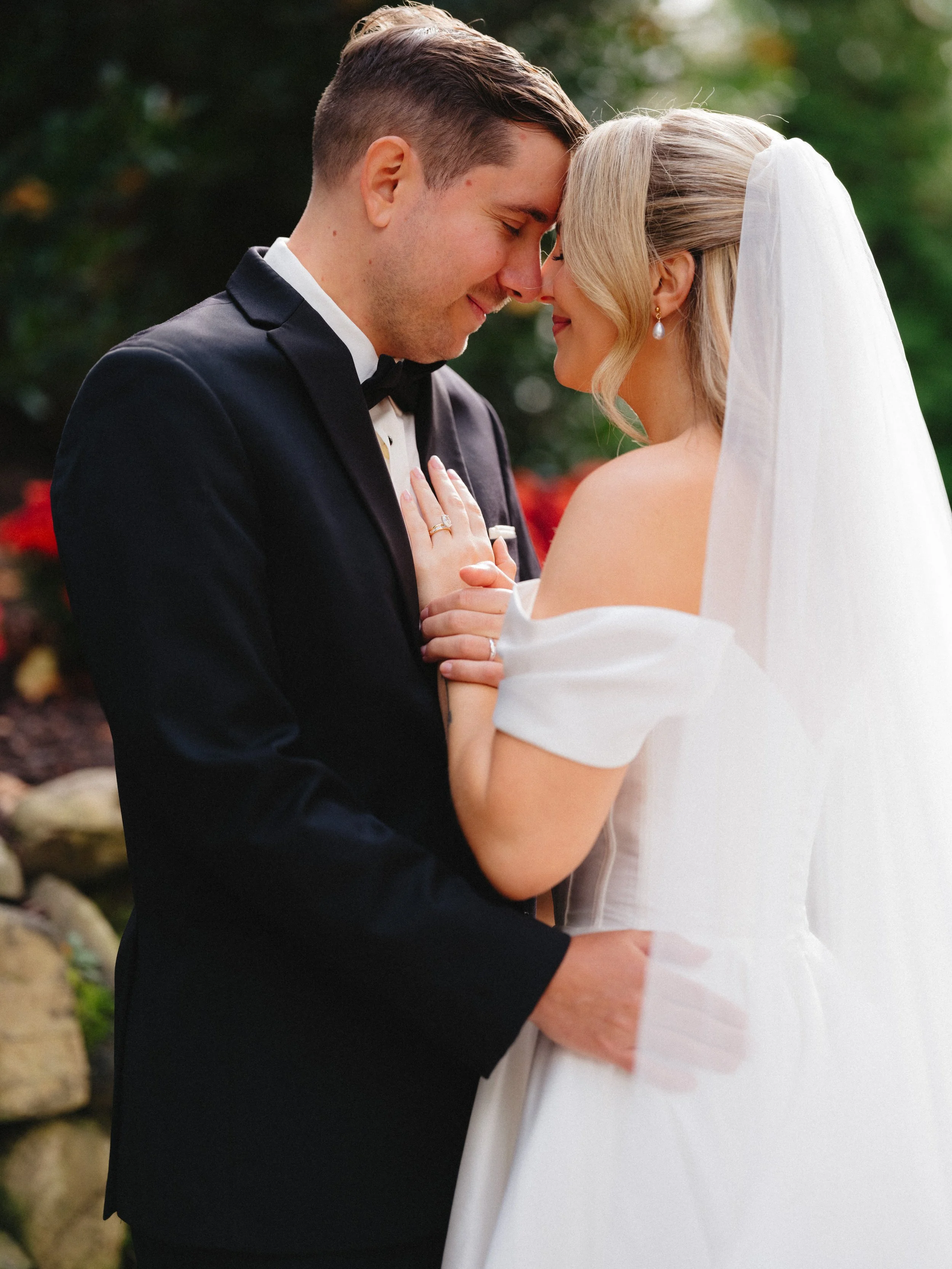 A bride and groom with foreheads touching, smiling outside during their wedding, with the bride wearing a white off-shoulder gown and veil, and the groom in a black tuxedo.