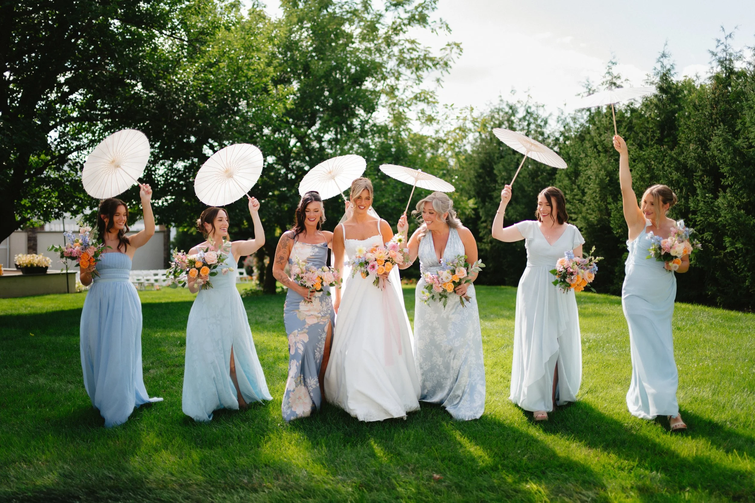 A bride and six bridesmaids outdoors, holding white parasols and bouquets, with greenery and trees in the background, on a sunny day.