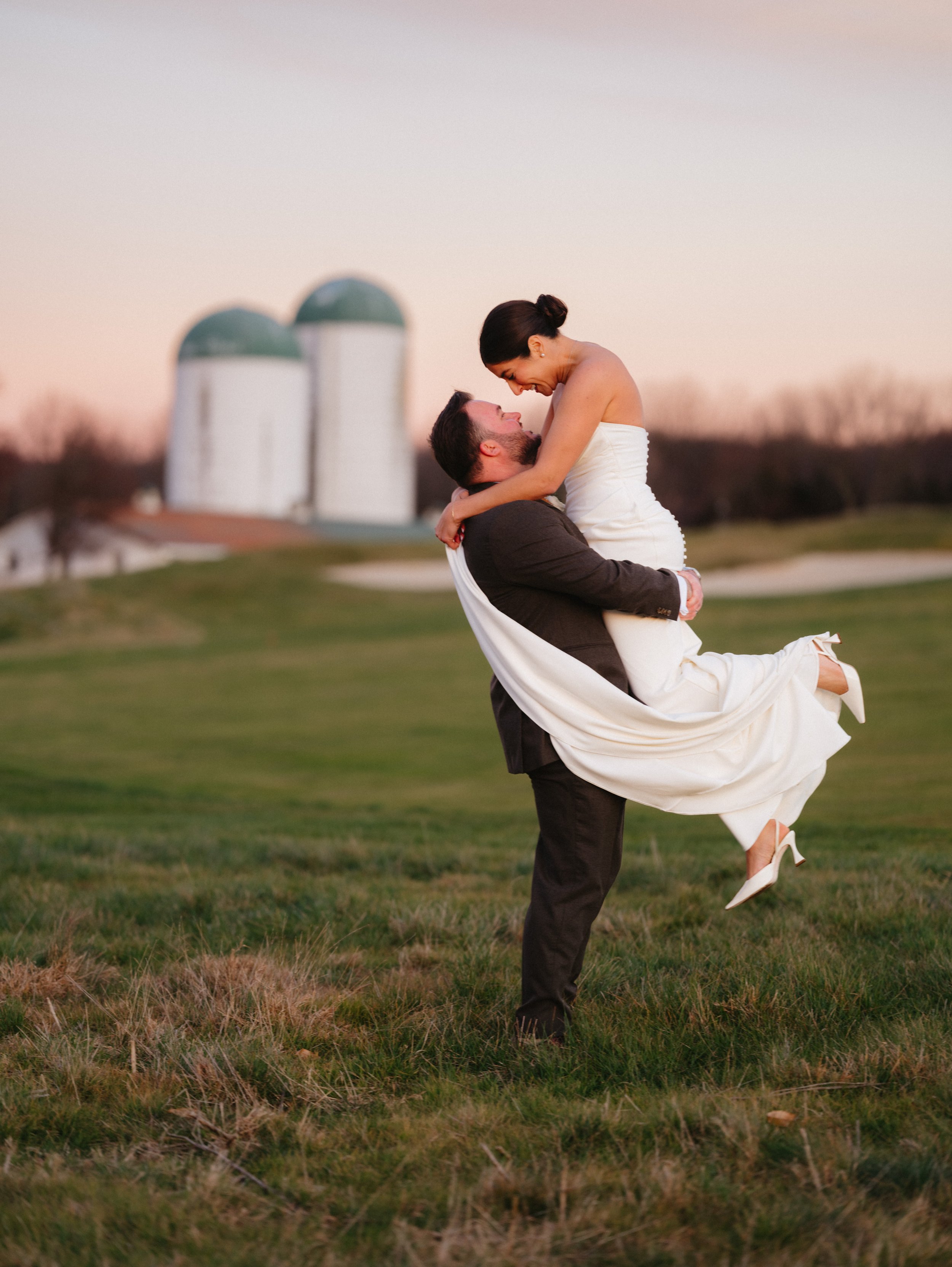 A man in a suit lifting a woman in a wedding dress in a field during sunset, with farm silos in the background.