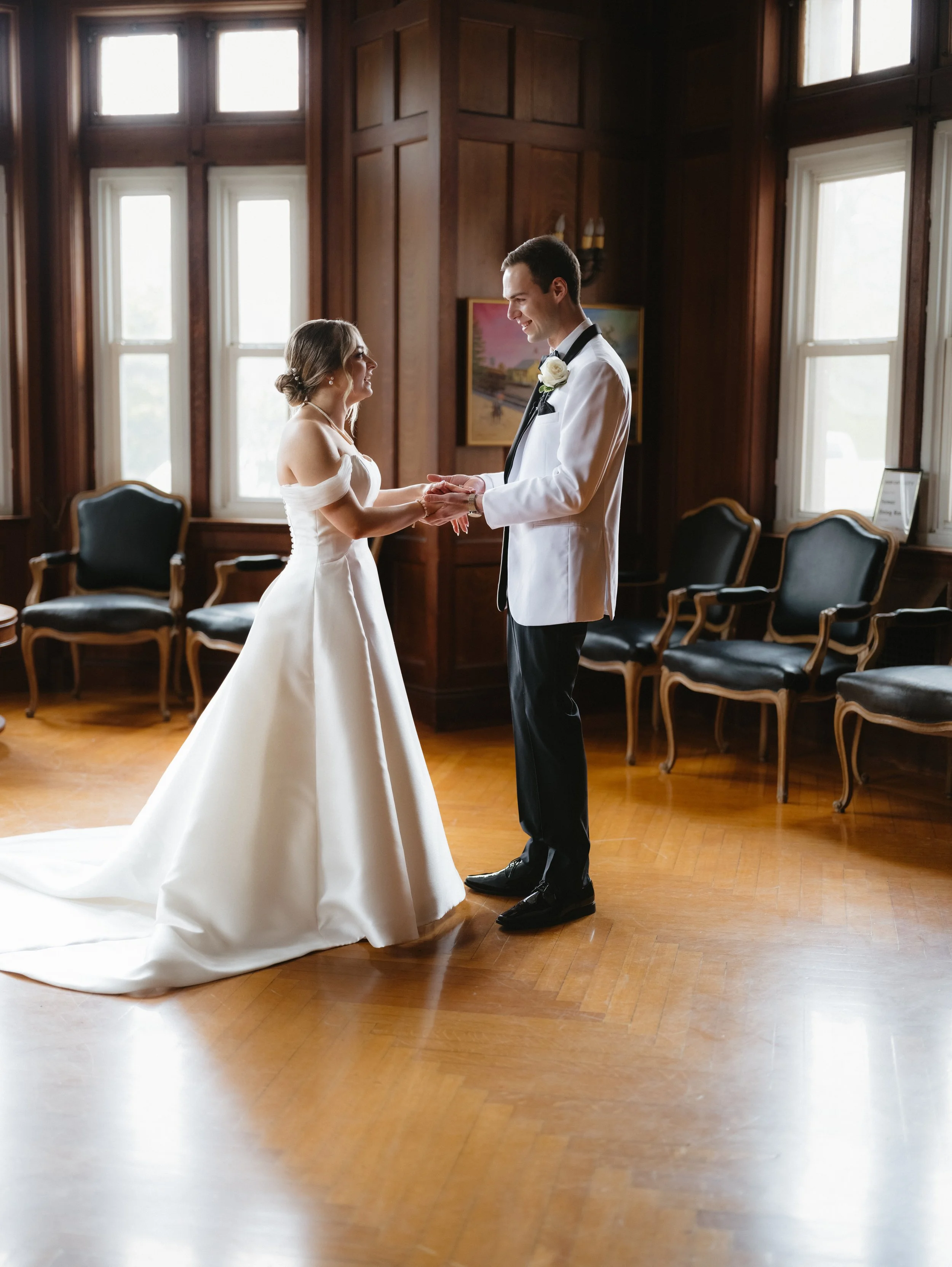 Bride and groom holding hands in a room with wooden floors and paneling, wearing a white wedding dress and a white tuxedo jacket respectively.