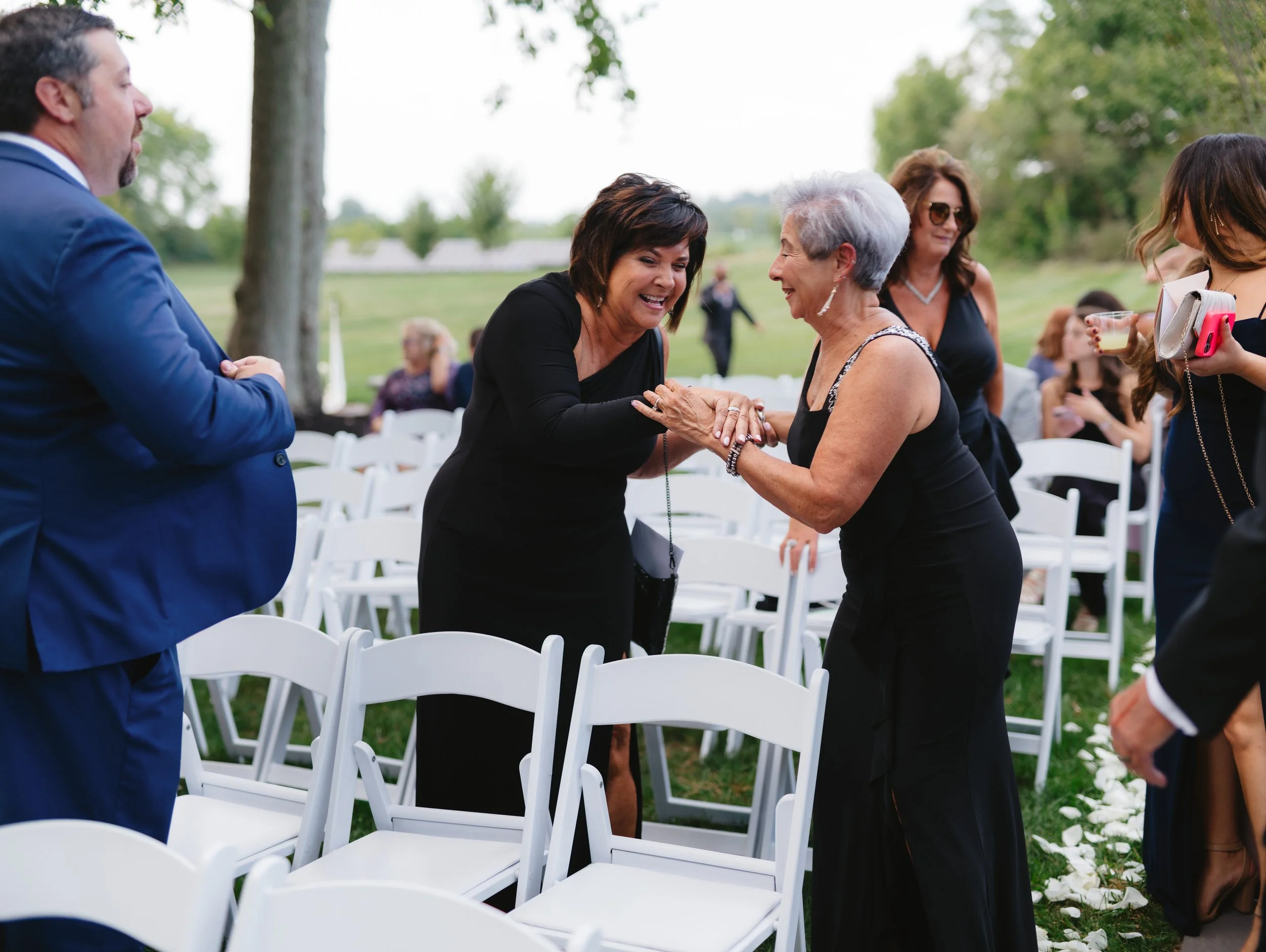 People in formal attire greet each other at an outdoor event with white chairs arranged in rows.
