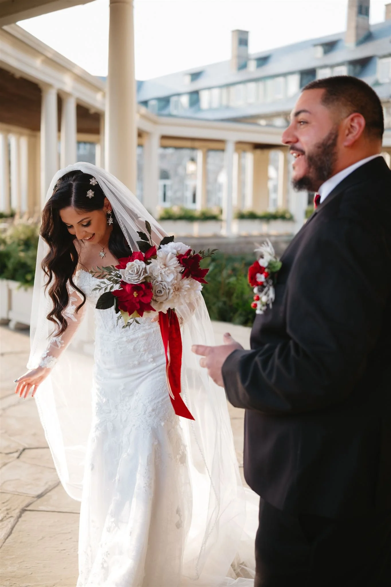 A bride and groom holding hands and walking outside, with the bride in a white gown and veil, and the groom in a black suit with a red boutonniere, in front of a building with stairs.