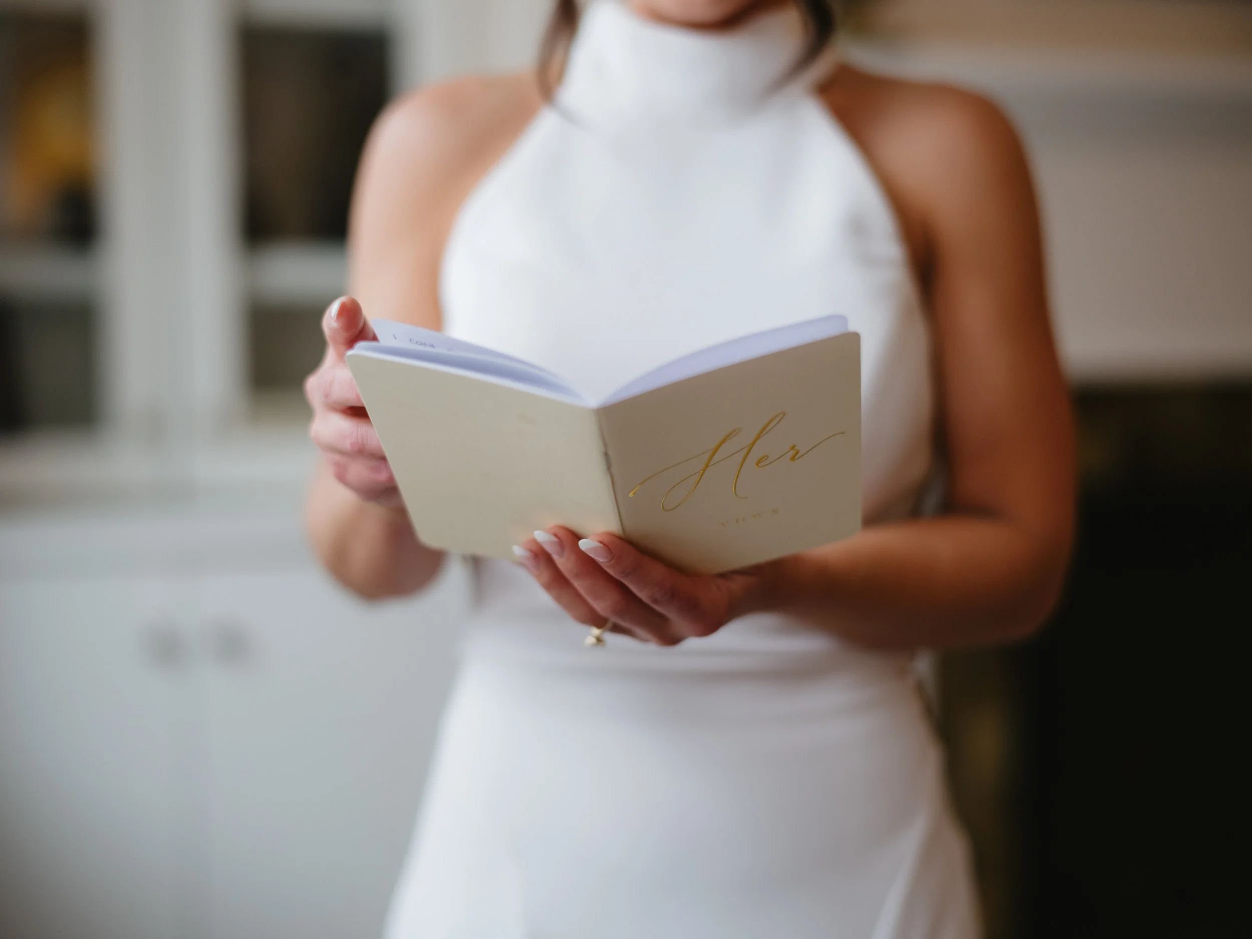 A woman in a white dress holding a booklet titled 'Her Vows' in an indoor setting.
