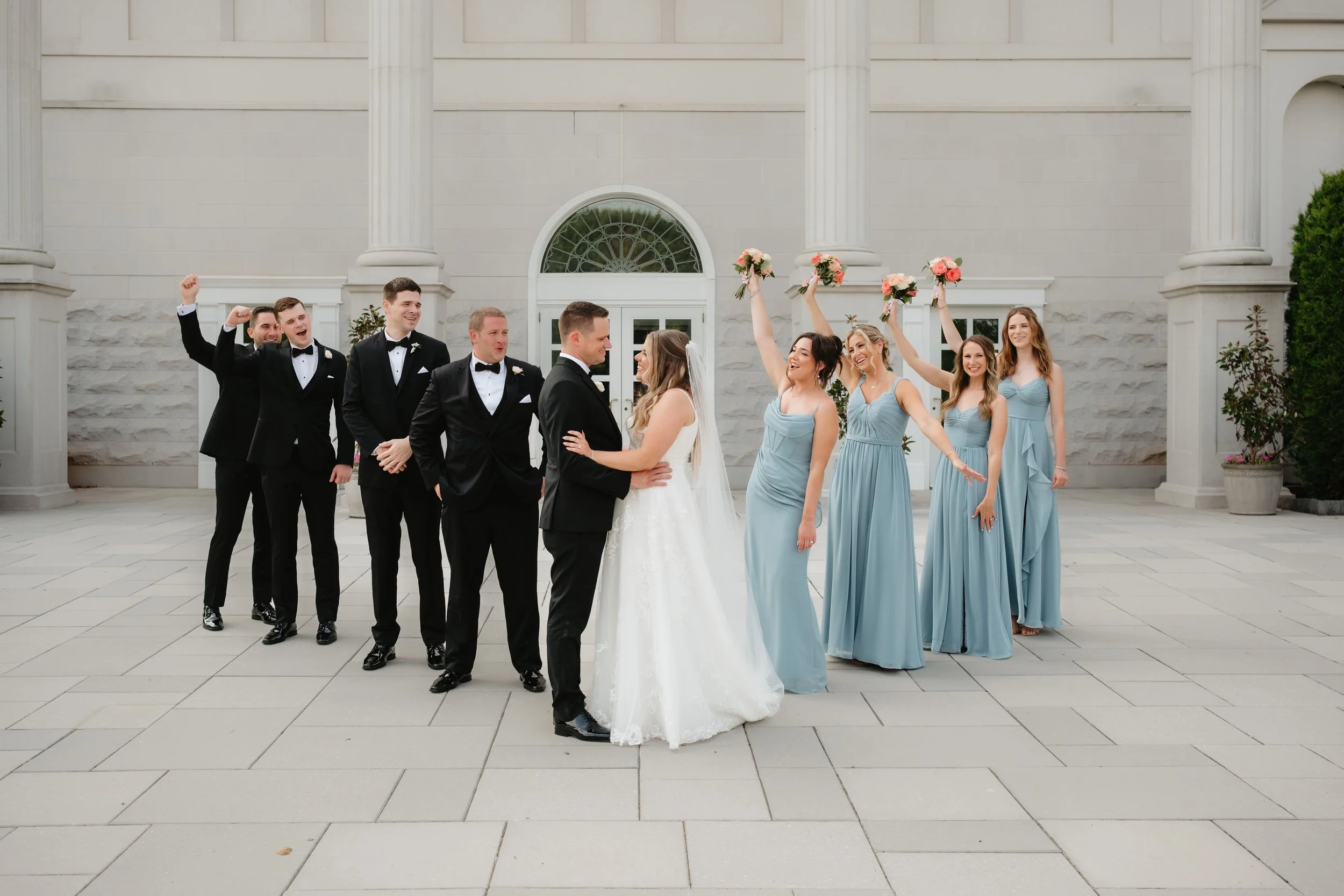 Wedding party photo with bride, groom, bridesmaids in light blue dresses, and groomsmen in black tuxedos.