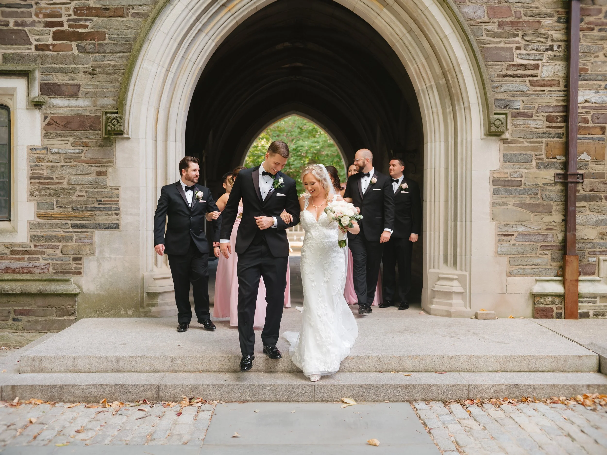 Wedding party walking through stone archway; bride in white lace gown holding a bouquet, groom in black tuxedo, accompanied by bridesmaids and groomsmen in formal attire. Stone building background.
