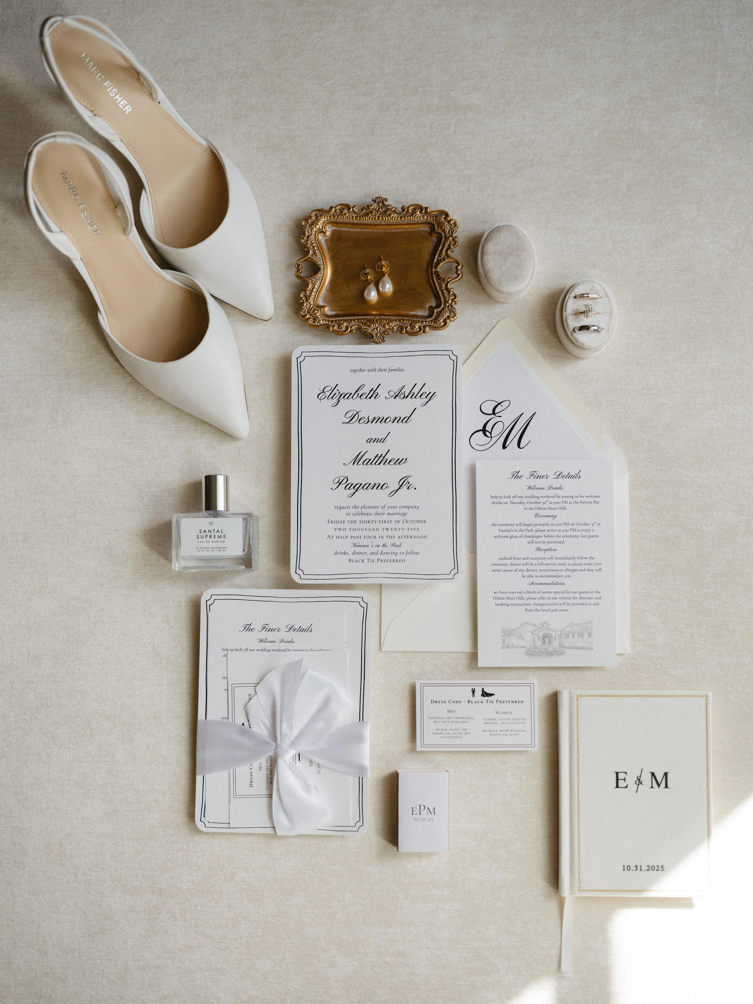 Wedding invitation suite with white high heels, pearl earrings, rings, perfume, and other wedding details on a cream-colored surface.