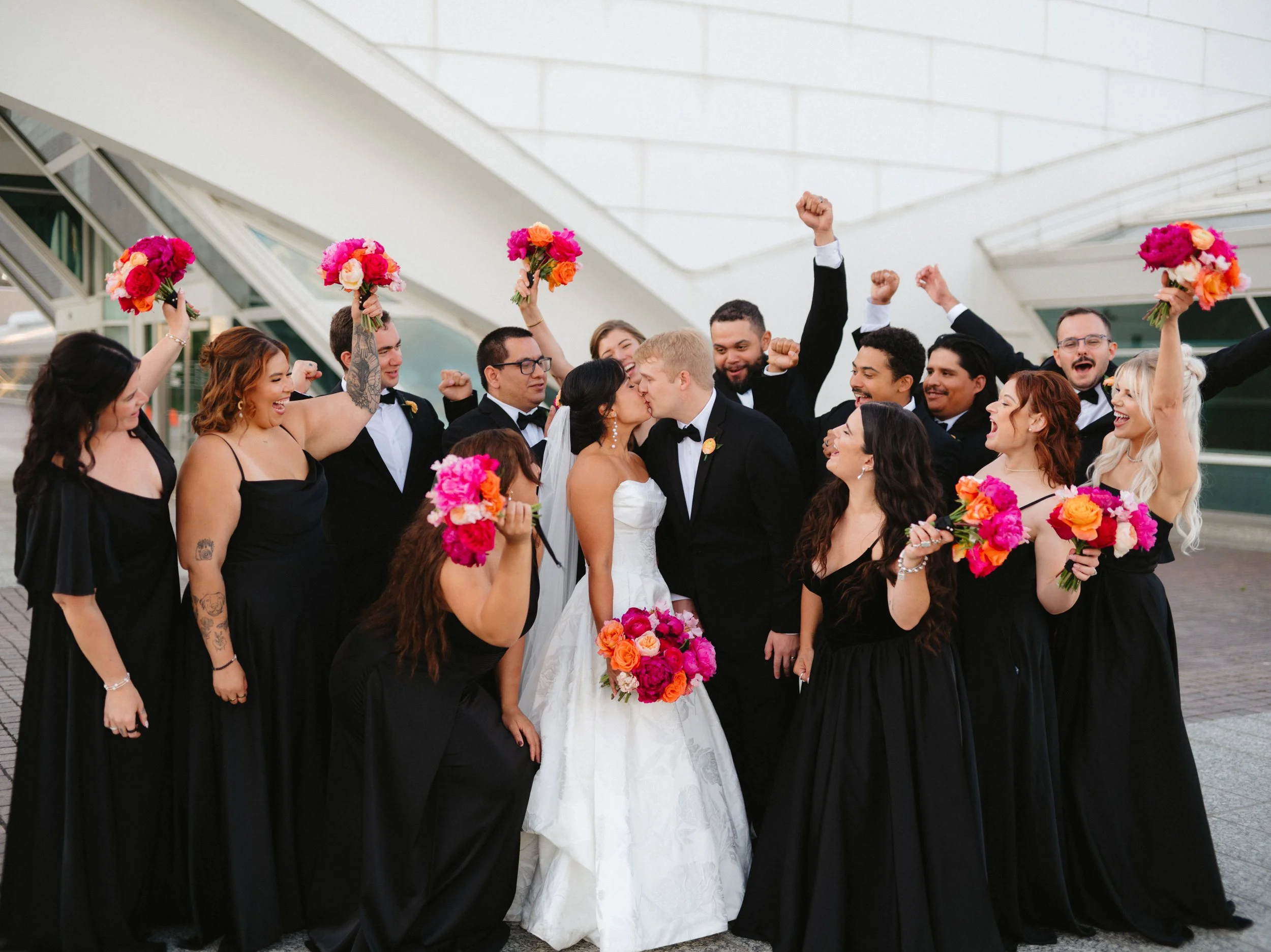 Wedding party celebrating on steps, bride and groom kissing, bridesmaids holding bouquets, groomsmen cheering.