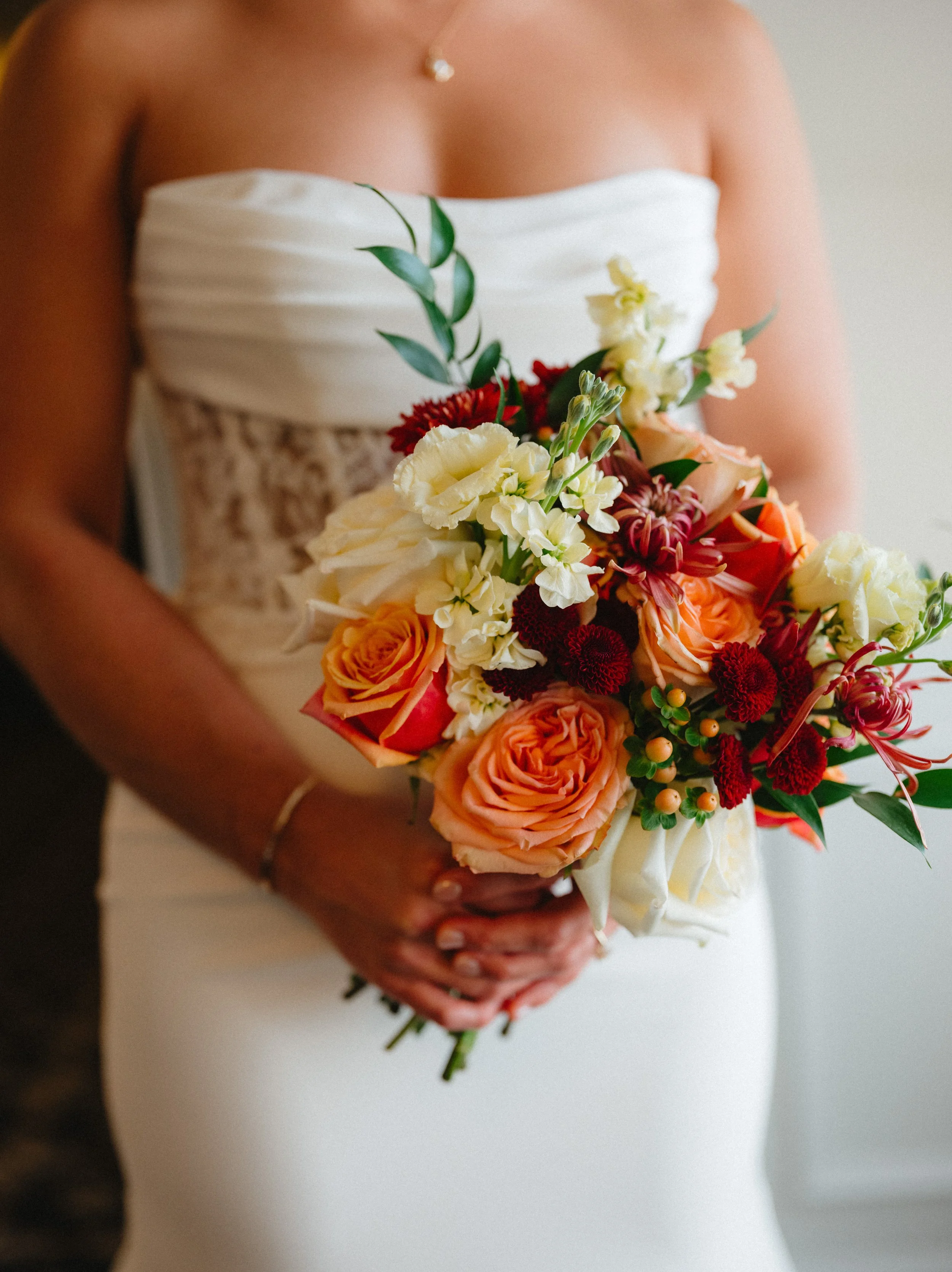 A bride holding a colorful bouquet of flowers including roses, dahlias, and greenery, in a strapless wedding dress.