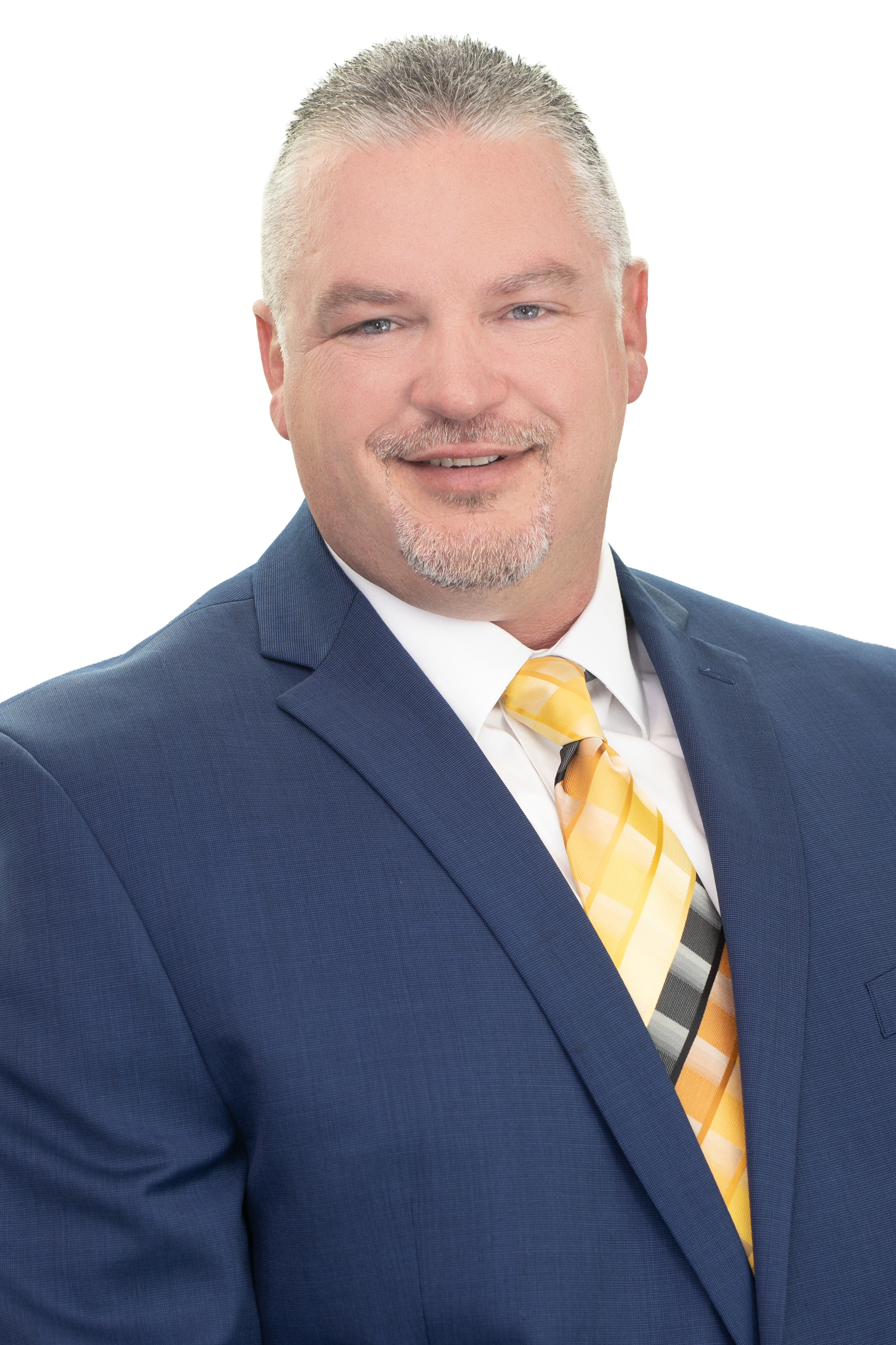 Head-and-shoulders professional portrait of a smiling man with short gray hair and a trimmed goatee, wearing a blue suit, white dress shirt, and yellow striped tie, seated indoors with a bright windowed background.