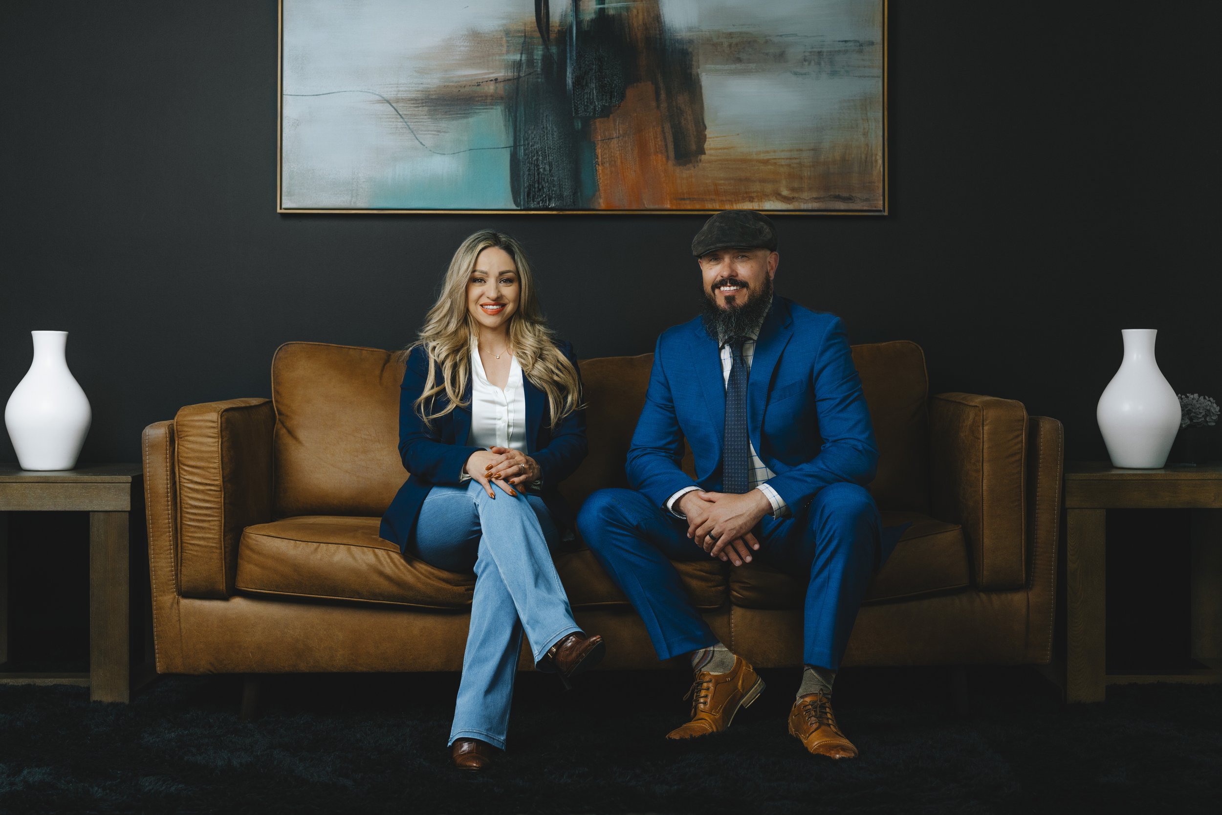 A woman and a man sitting on a brown leather sofa in a modern room, dressed in business attire. The background features dark walls, a large abstract painting, and two white vases on side tables.