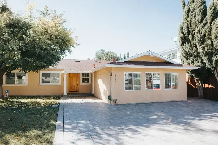 Suburban house with a yellow exterior, large front windows, wooden front door, paved driveway, and surrounding trees with garage conversion.