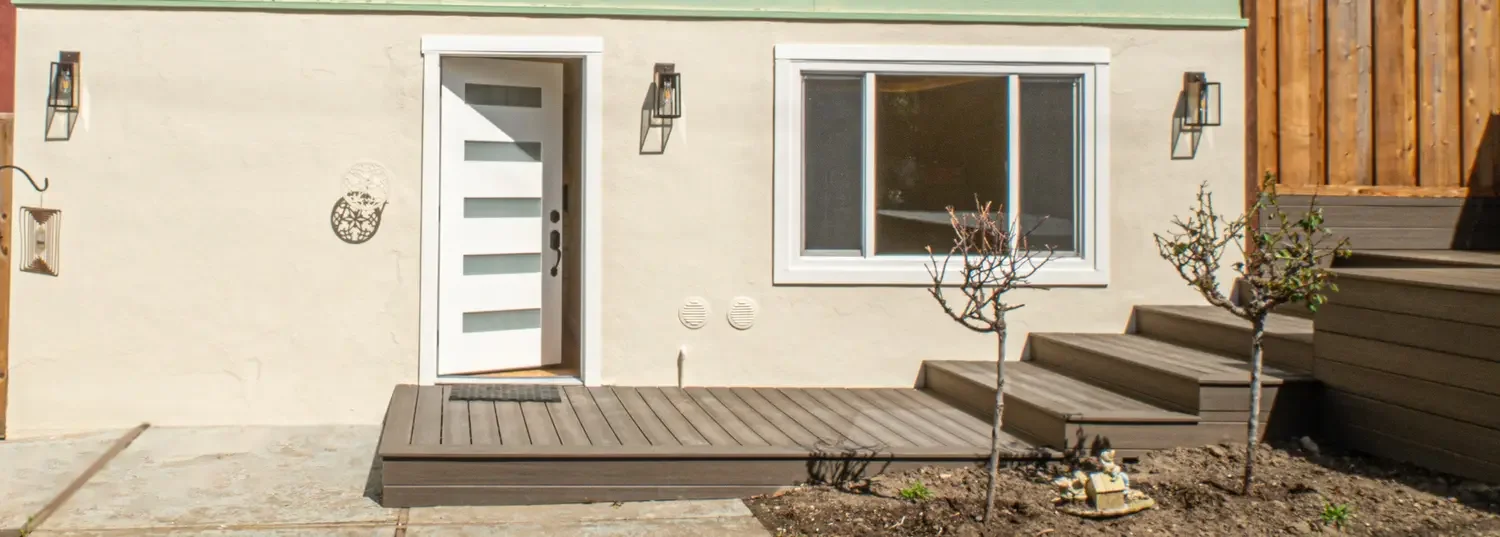 Exterior of Modern 1 bedroom home conversion exterior with a white front door, glass panels, adjacent window, and wooden steps. Minimalist landscaping and wall-mounted lanterns.