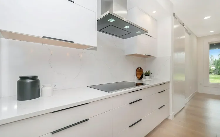 Modern ADU kitchen with white cabinets, marble backsplash, black induction cooktop, and stainless steel range hood. Two decorative jars, a cutting board, and potted plant on counter.