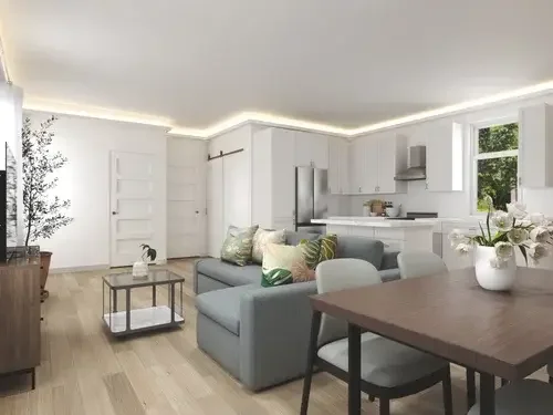 Open living room and kitchen with white cabinets, a gray sofa with decorative pillows, a wooden dining table with a white vase of flowers, and large window showing greenery outside.