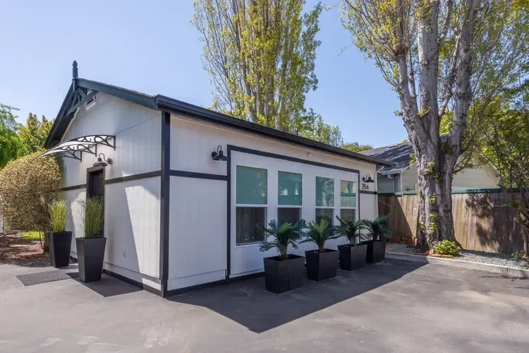 Detached Garage Conversion one bedroom ADU with black trim, large windows, and potted plants in the driveway next to tall trees.