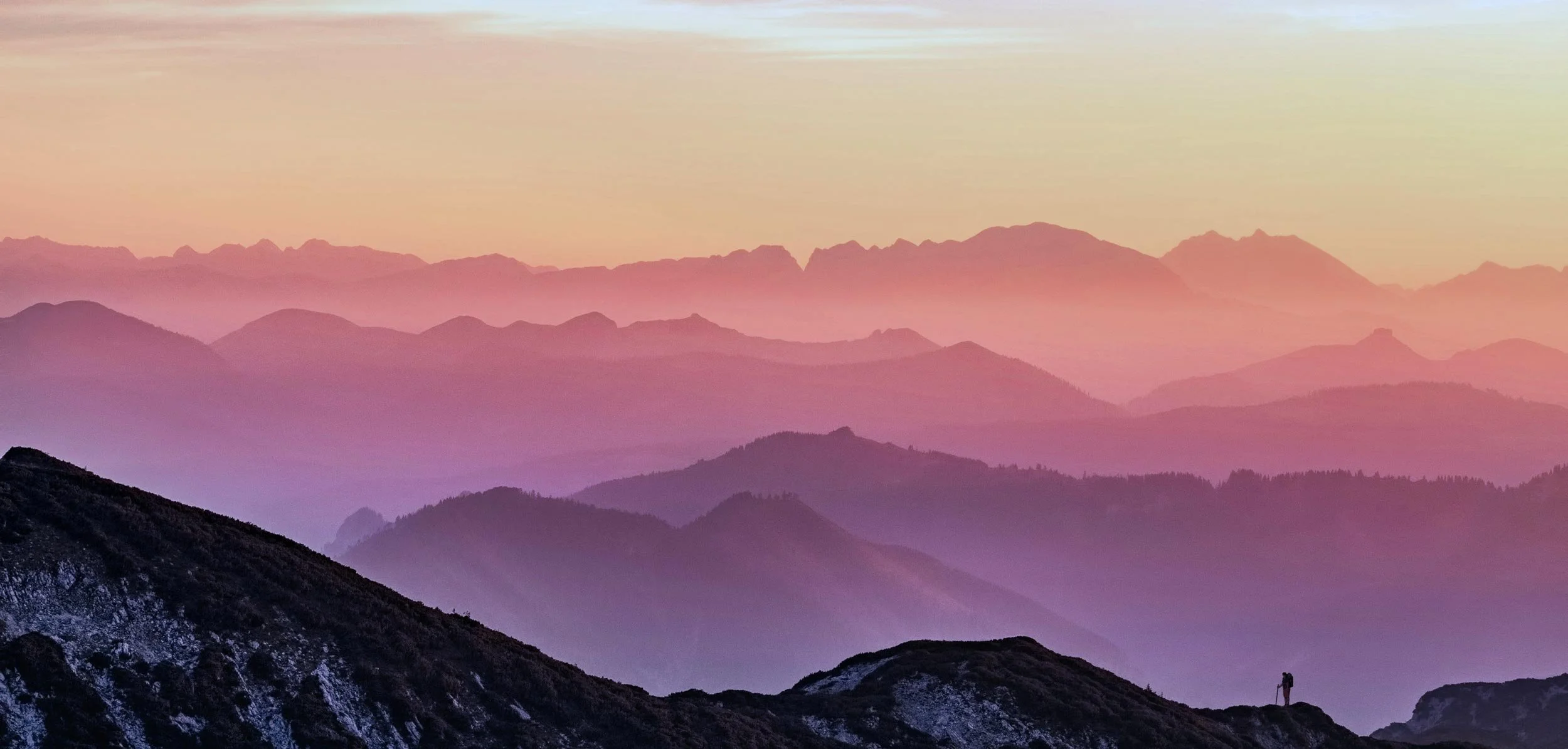 Mountain landscape at dusk with one solo hiker on a peak, purple orange and pink tones accentuate the colours of the sunset.