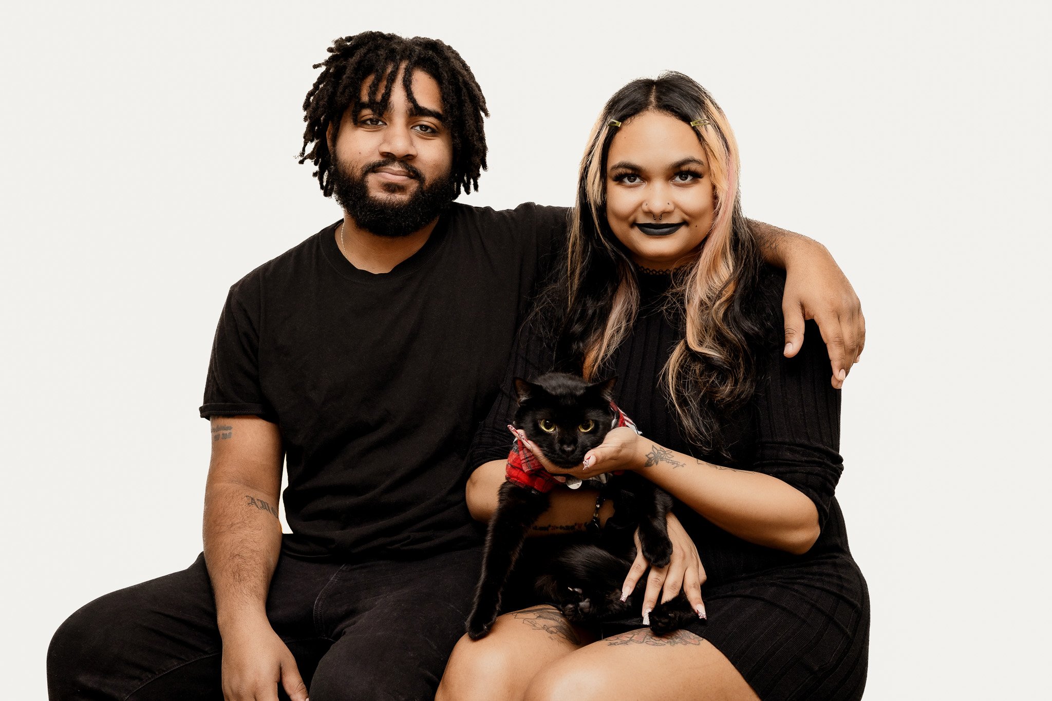 A diverse young couple sitting closely together against a plain white background. The woman has long, two-toned hair with blonde and black streaks, wears black lipstick, and is holding a black cat dressed in a red plaid outfit. The man has shoulder-l