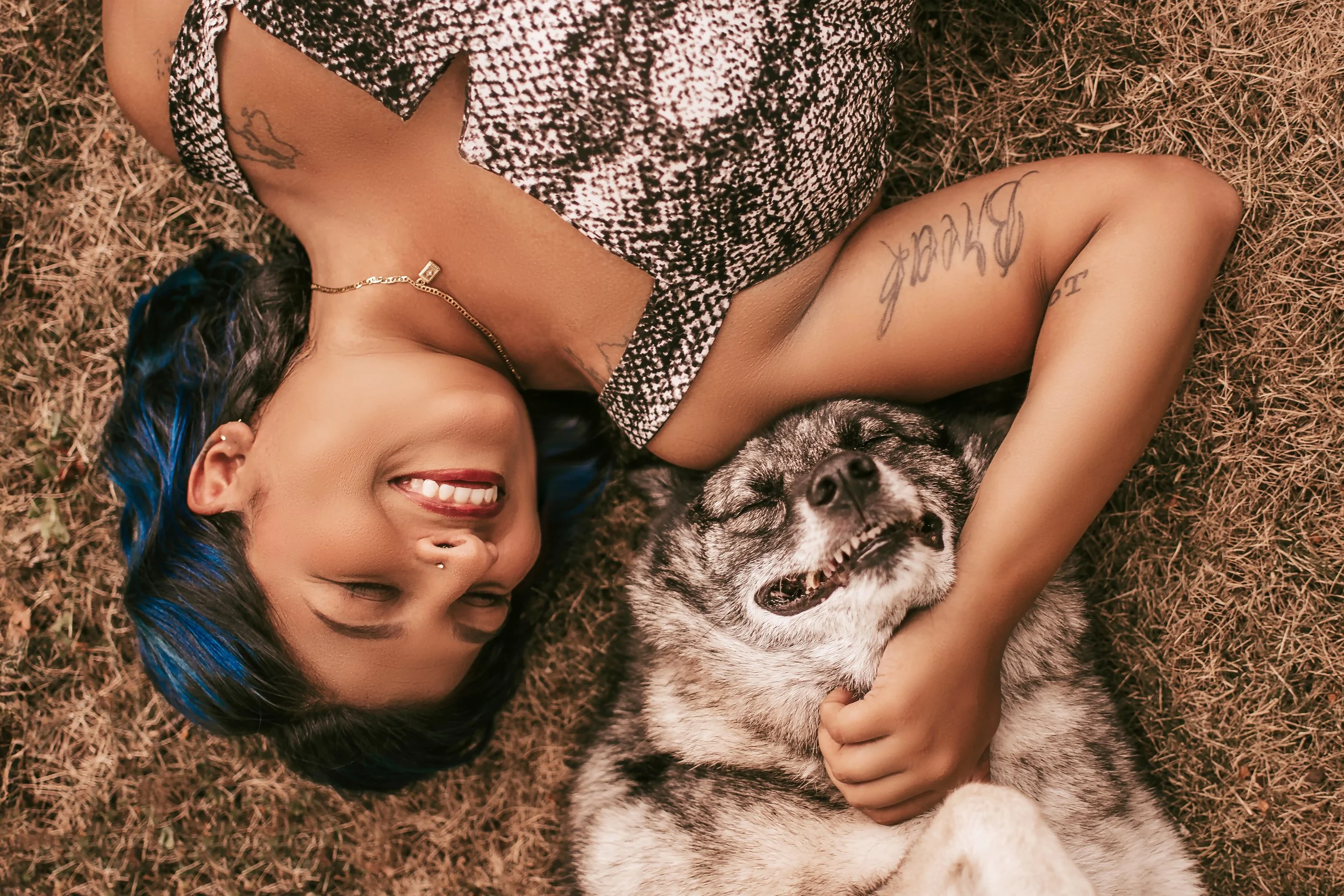 A young woman with dark hair lying on the ground with a Husky dog, smiling and holding the dog, outdoors on dry grass.