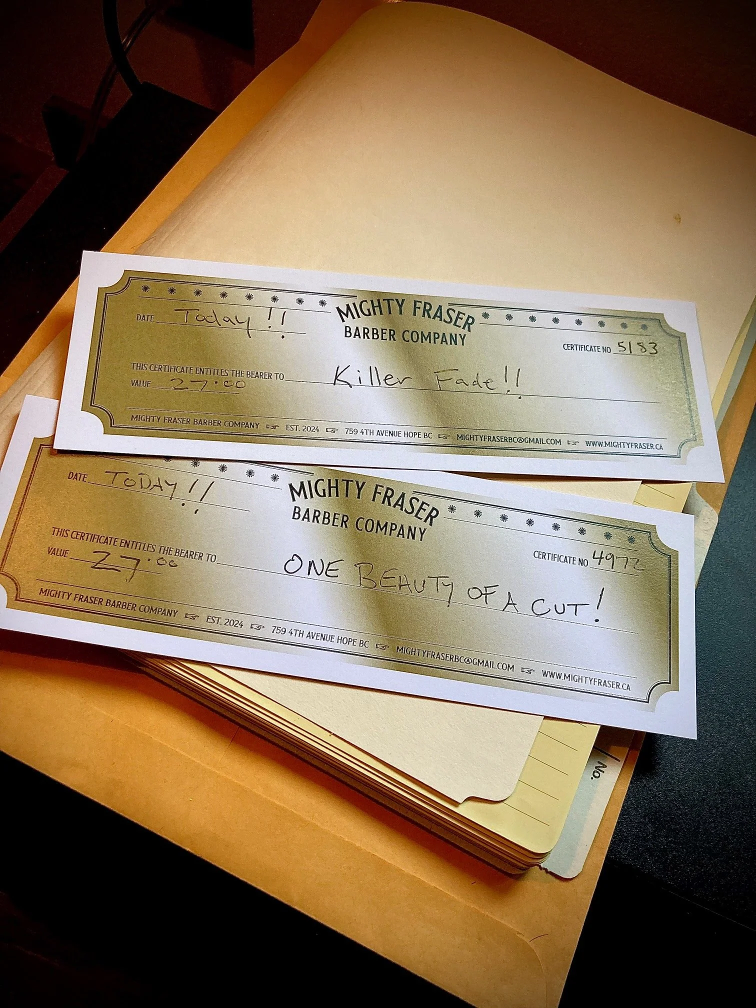 Two barber gift certificates from Mighty Fraser Barber Company, one for a haircut called Killer Fade and the other for a haircut called One Beauty of a Cut, placed on top of a stack of papers on a wooden desk.
