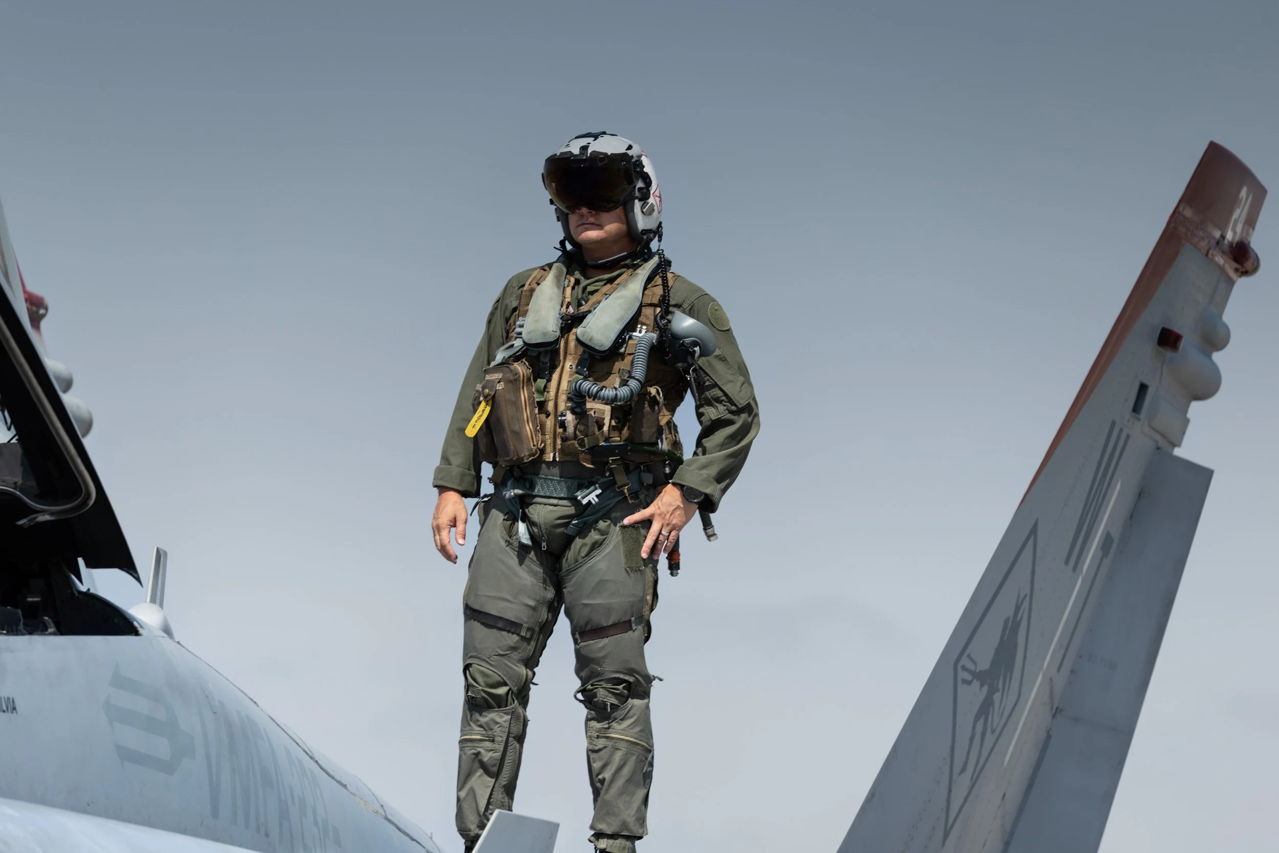 A pilot in military flight gear standing on the wing of an aircraft, wearing a helmet, flight suit, and harness, with the sky in the background.