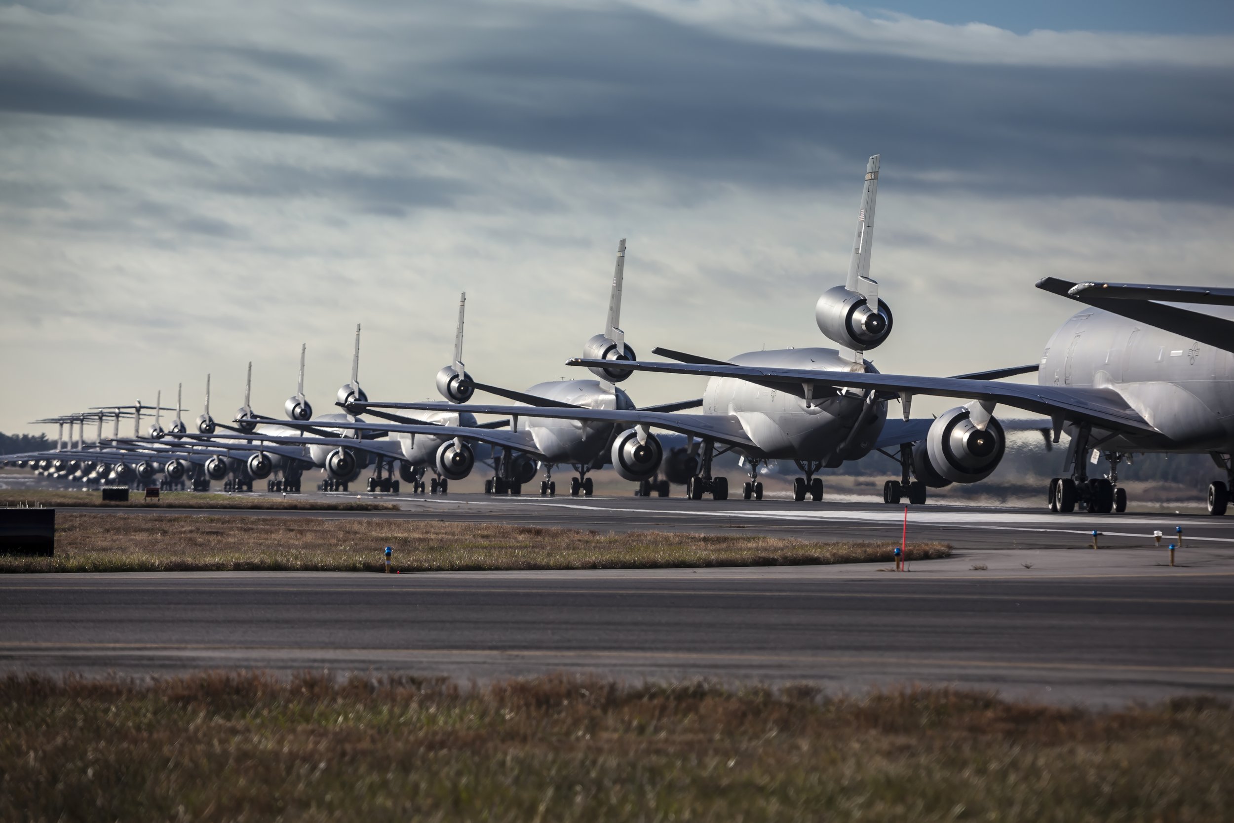 Multiple military transport aircraft lined up on an airport runway under a cloudy sky.