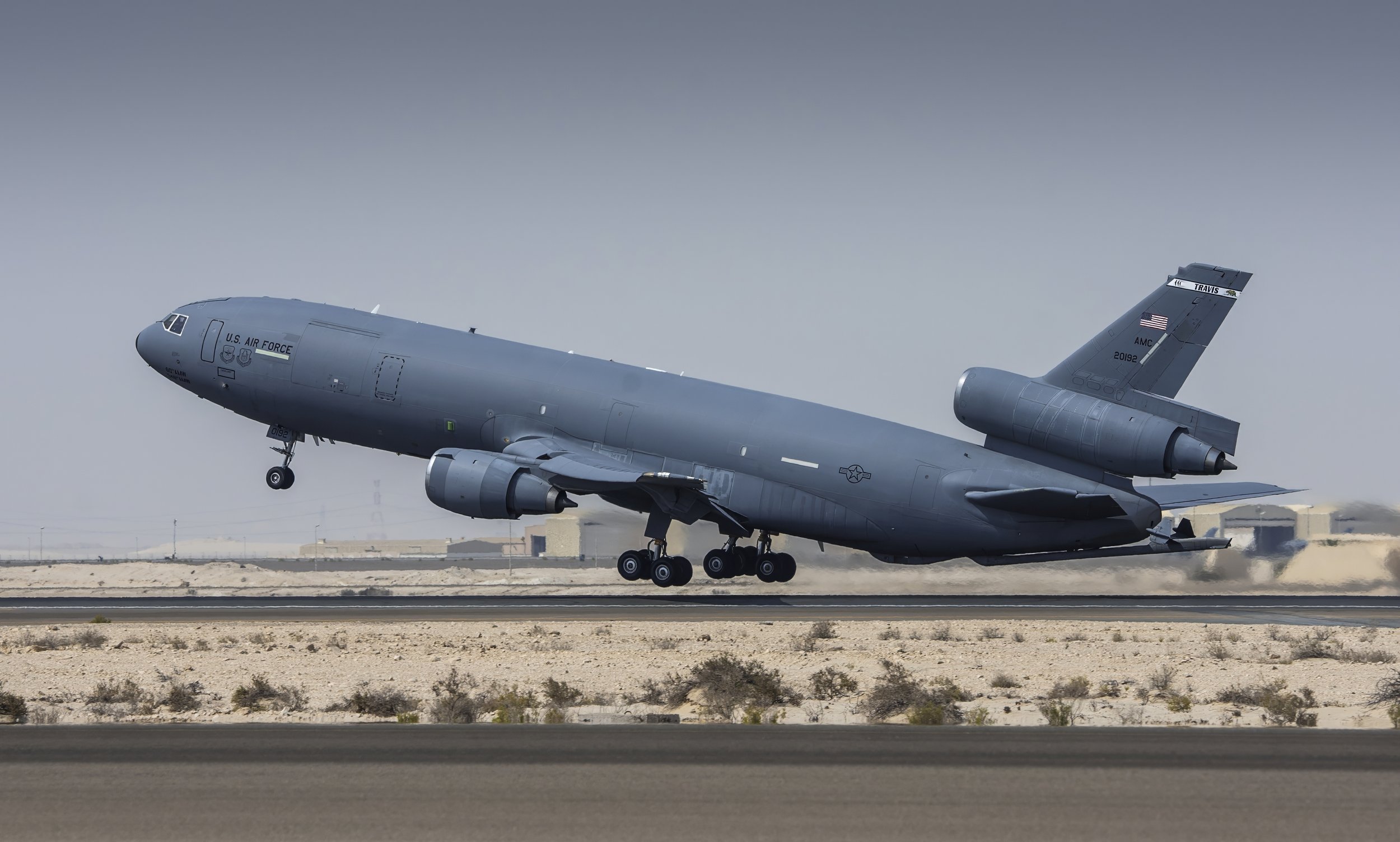 A gray U.S. Air Force aircraft taking off from a desert runway with housing structures in the background.
