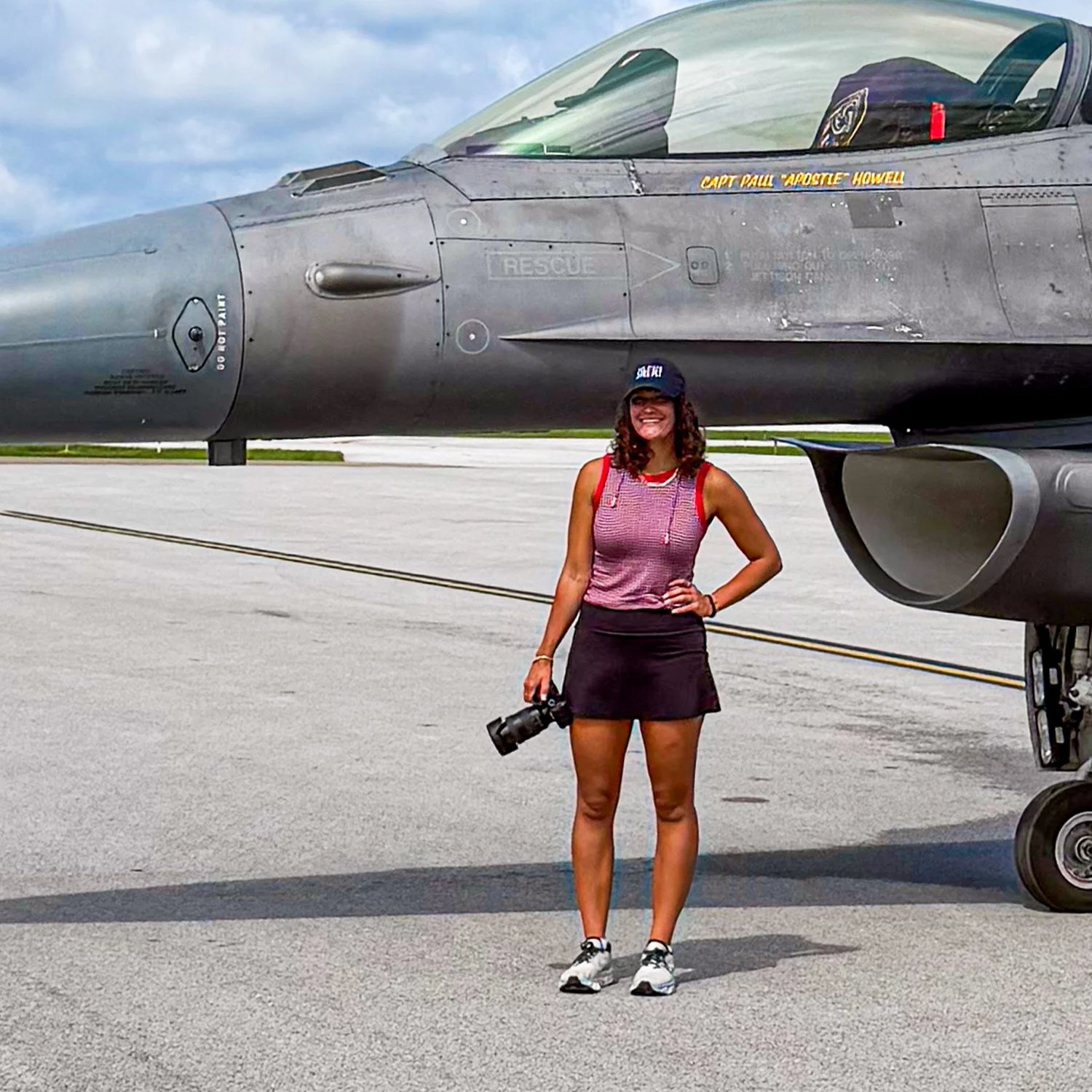 A woman smiling and holding a camera stands in front of a fighter jet aircraft on an airfield. The woman is wearing a red and white tank top, a black skirt, and a navy blue cap.
