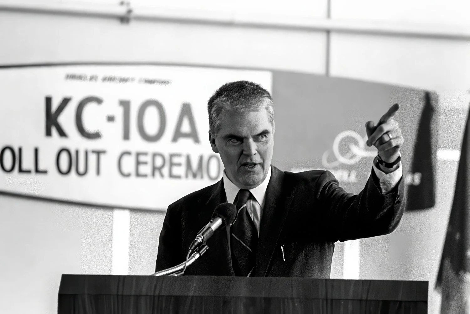 A man in a suit and tie speaking at a podium with a serious expression, pointing with his right hand, in front of a sign that reads "KC-10A ROLL OUT CEREMO".