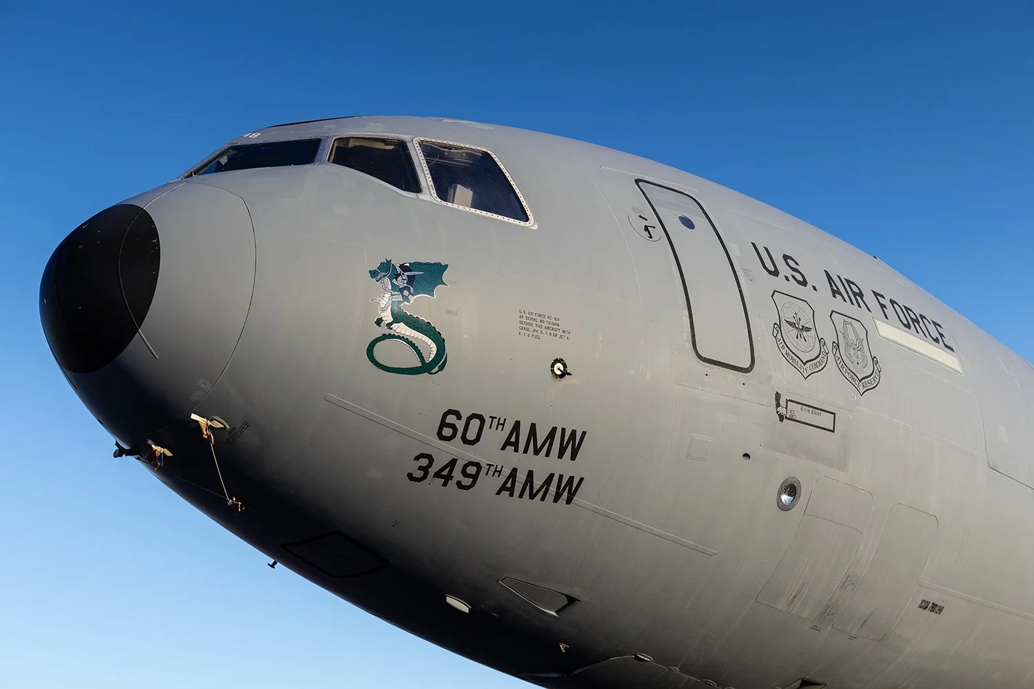 Close-up of a U.S. Air Force aircraft nose with military insignias and markings, including a dragon emblem and text indicating it belongs to the 60th and 349th Air Mobility Wings, against a clear blue sky.