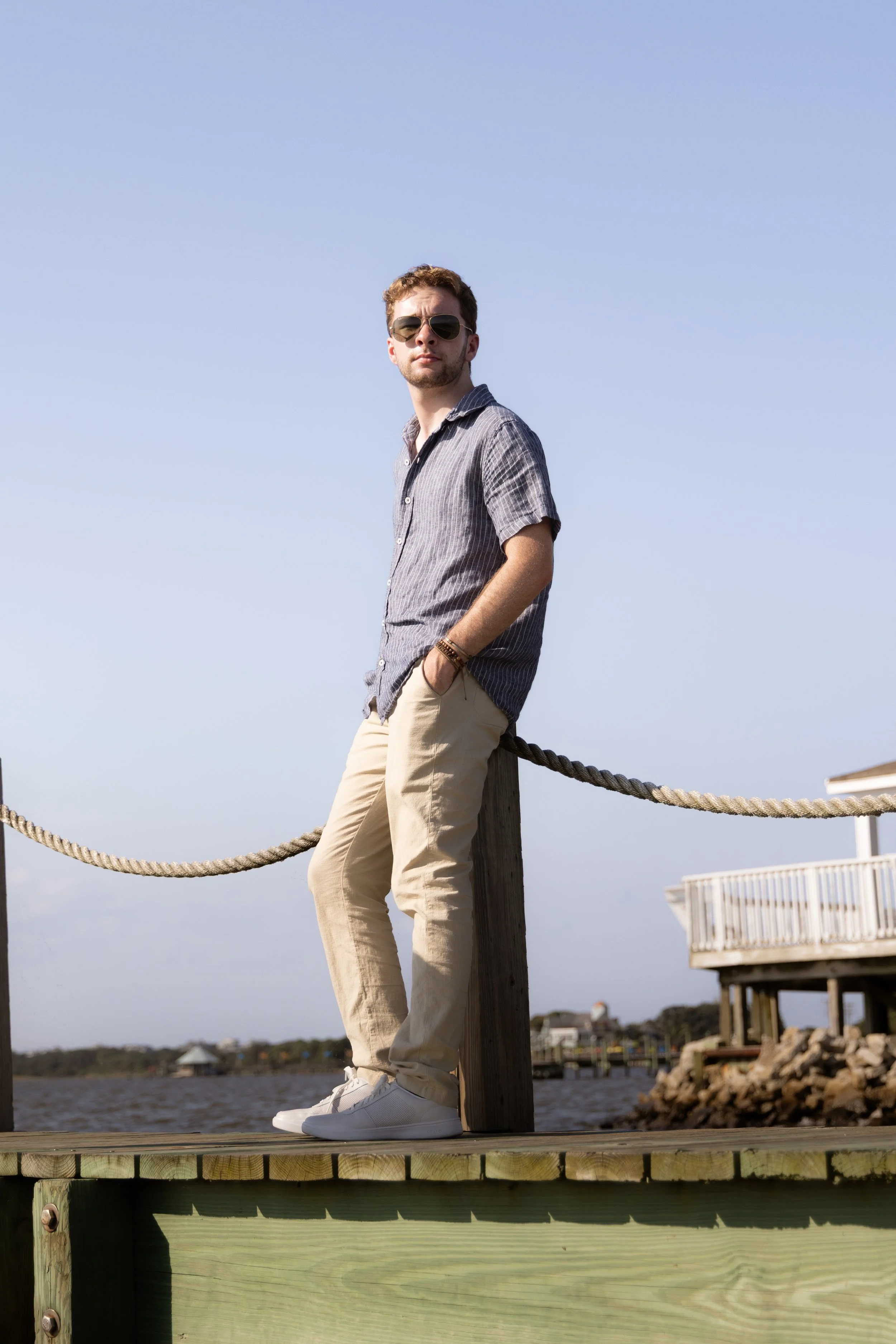 A man with sunglasses and casual clothing leaning against a wooden post on a dock near the water, with houses and a blue sky in the background.