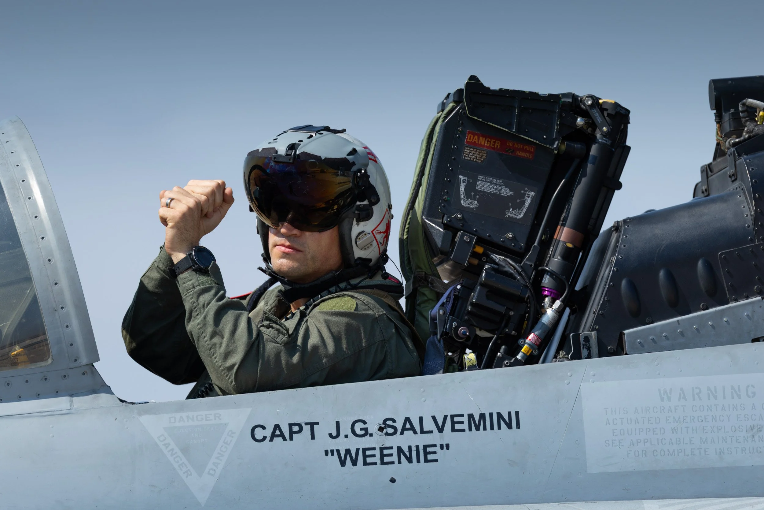 A fighter pilot sitting in the cockpit of a military aircraft, wearing a helmet and sunglasses, with one arm flexed. The aircraft has a label indicating the pilot's name as Captain J.G. Salvemini, nicknamed "Weenie."