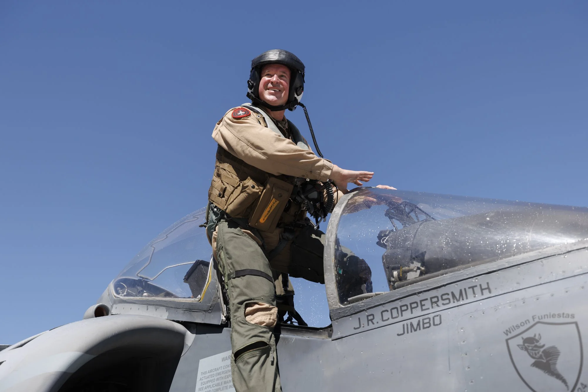 A smiling man in pilot gear standing next to an aircraft cockpit, wearing a helmet and a tan flight suit.