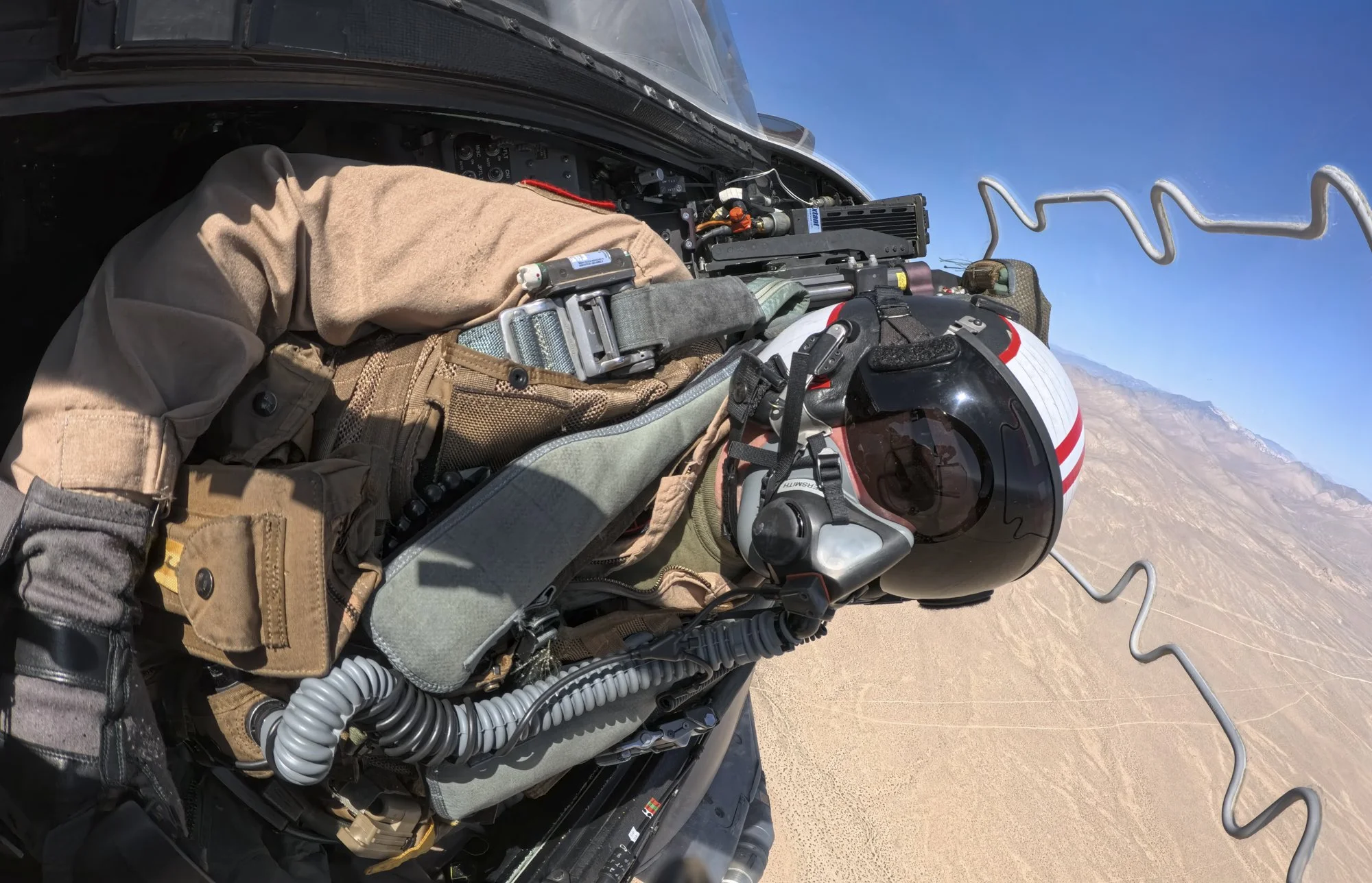 A military pilot in a tan uniform and helmet with red and white stripes flying a fighter jet over a desert landscape with mountains in the distance.