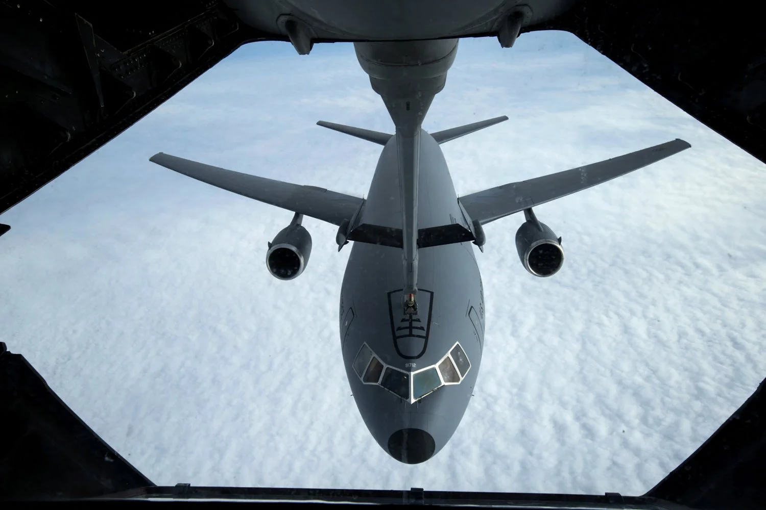An aerial view of a fighter jet seen from inside the cargo bay of an aircraft, with the sky and clouds below.