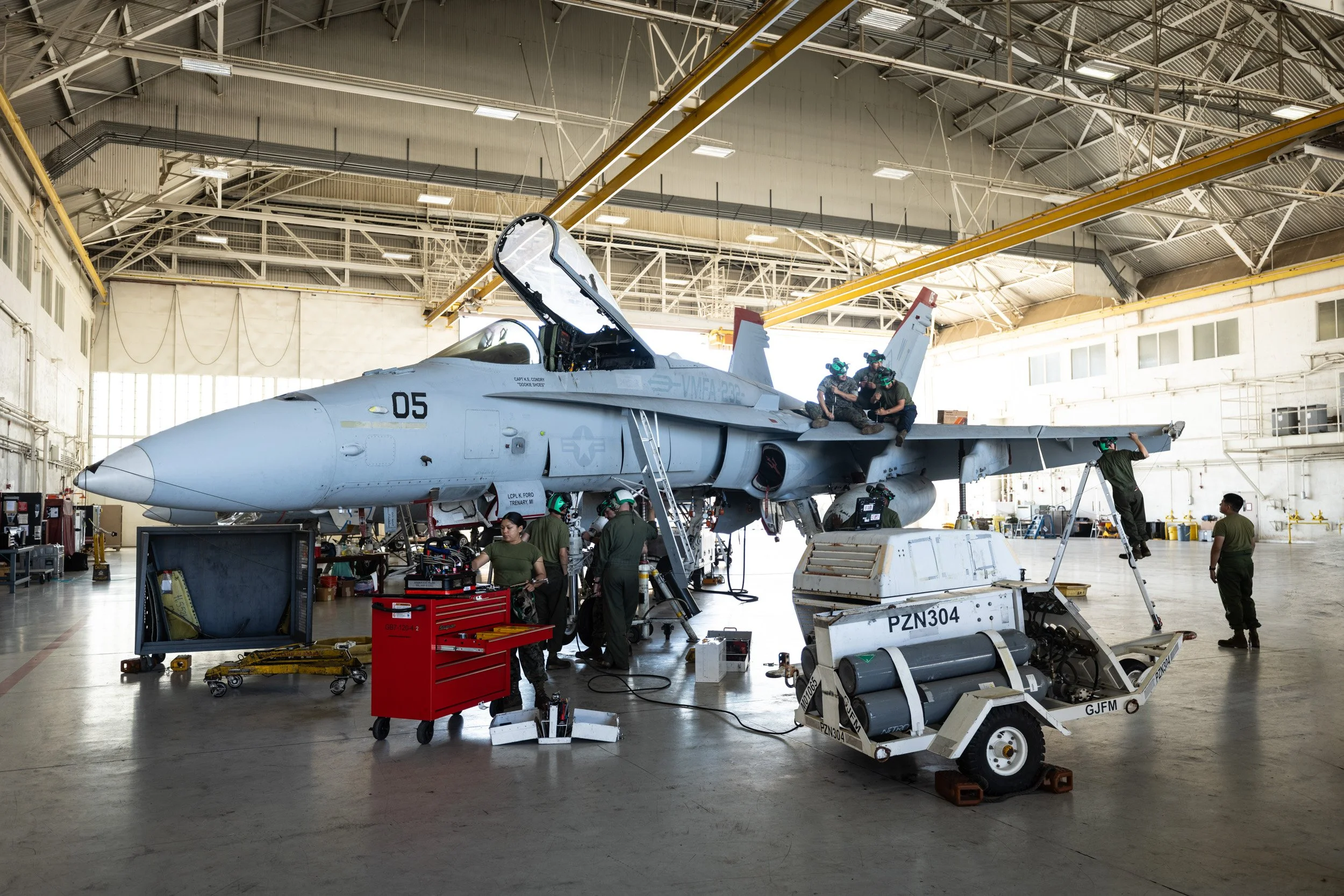 U.S. Navy fighter jet inside an aircraft hangar undergoing maintenance, with personnel working around it and equipment nearby.