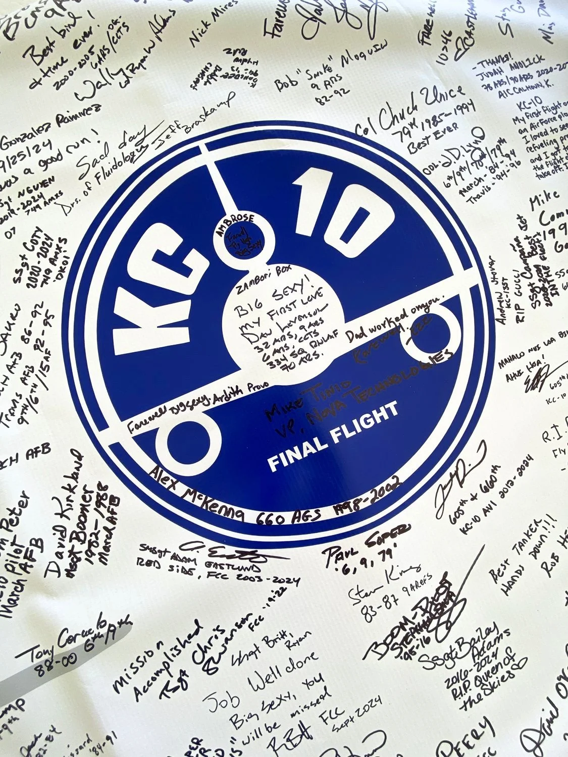 A circular blue sign with white text reading "VFC 10" and "FINAL FLIGHT." It contains numerous handwritten notes and signatures surrounding the sign, with some names, dates, and messages, including farewell messages, signatures, and personal notes.