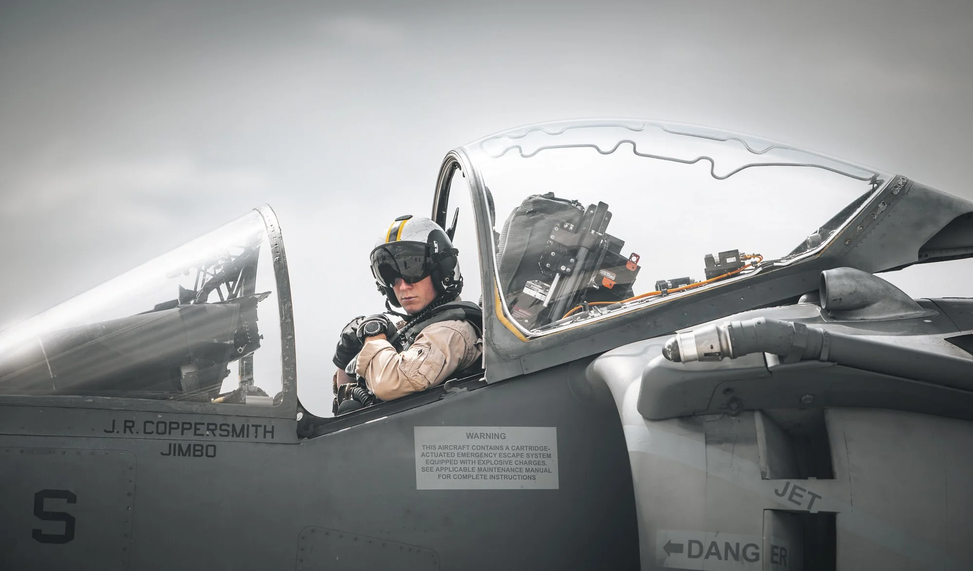 Pilot sitting in the cockpit of a fighter jet, wearing a helmet with a visor and beige flight suit, with the jet's canopy open.