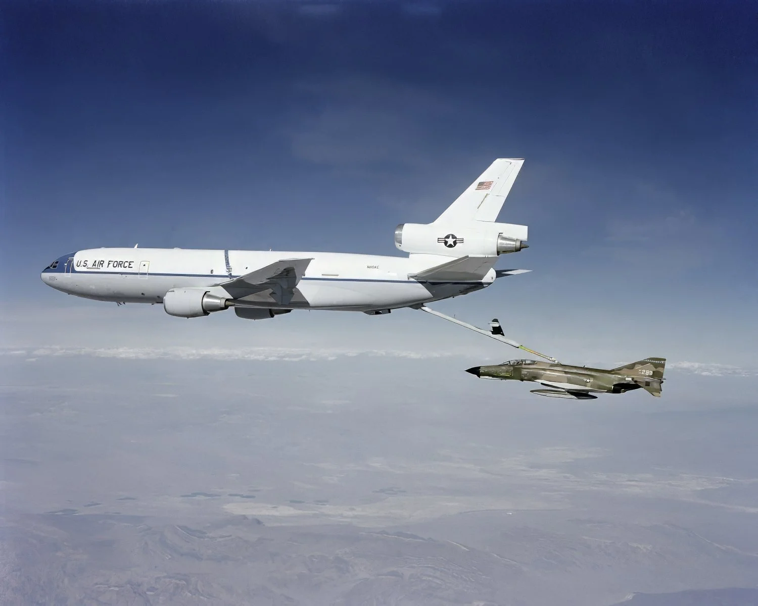 A U.S. Air Force Boeing KC-135 Stratotanker aircraft refueling a military fighter jet mid-air against a clear sky and cloudy landscape below.