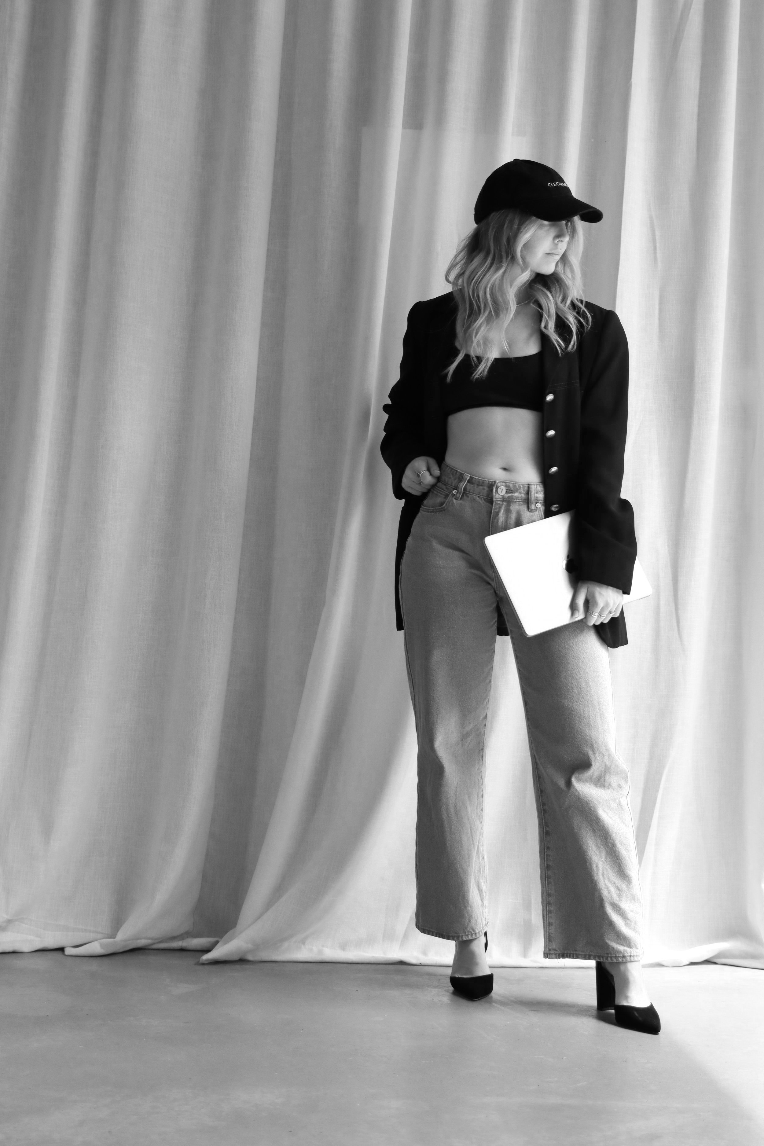 A woman stands in front of a curtain, wearing a black cap, black cropped top, open blazer, wide-leg jeans, and high heels, holding a laptop.