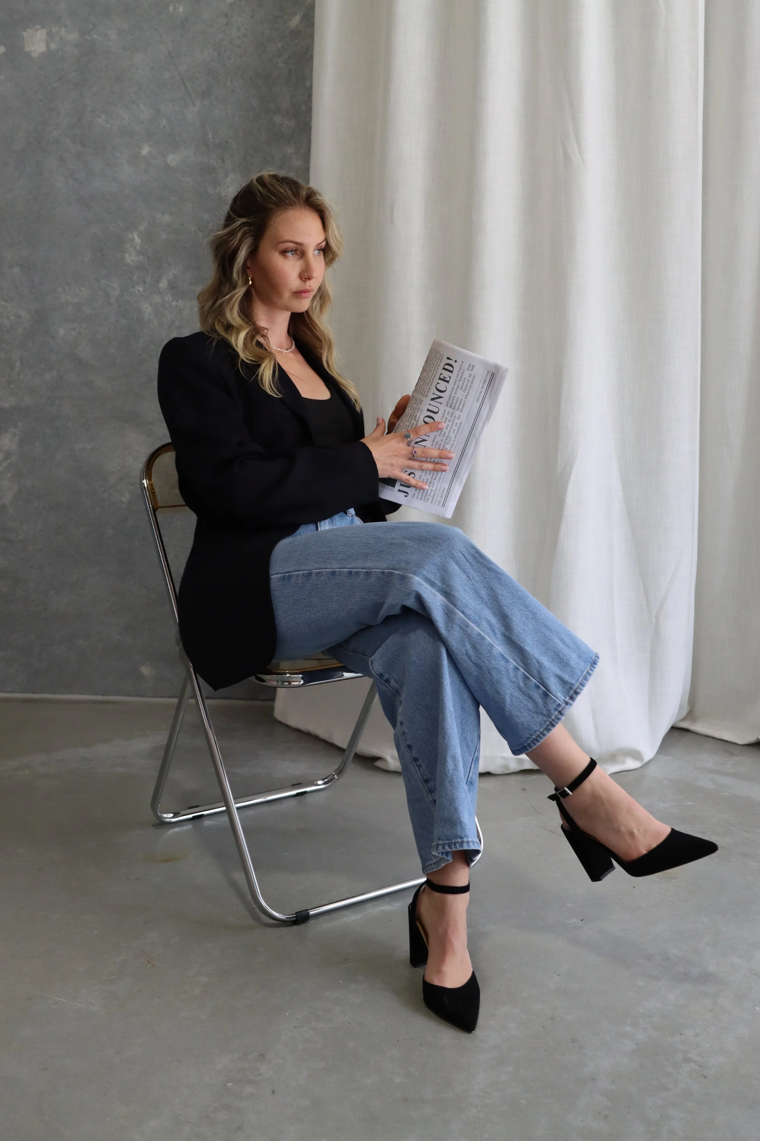 A woman with blonde hair, wearing a black blazer, jeans, and black heels, sitting on a metal chair, reading a newspaper indoors near white curtains and a gray wall.