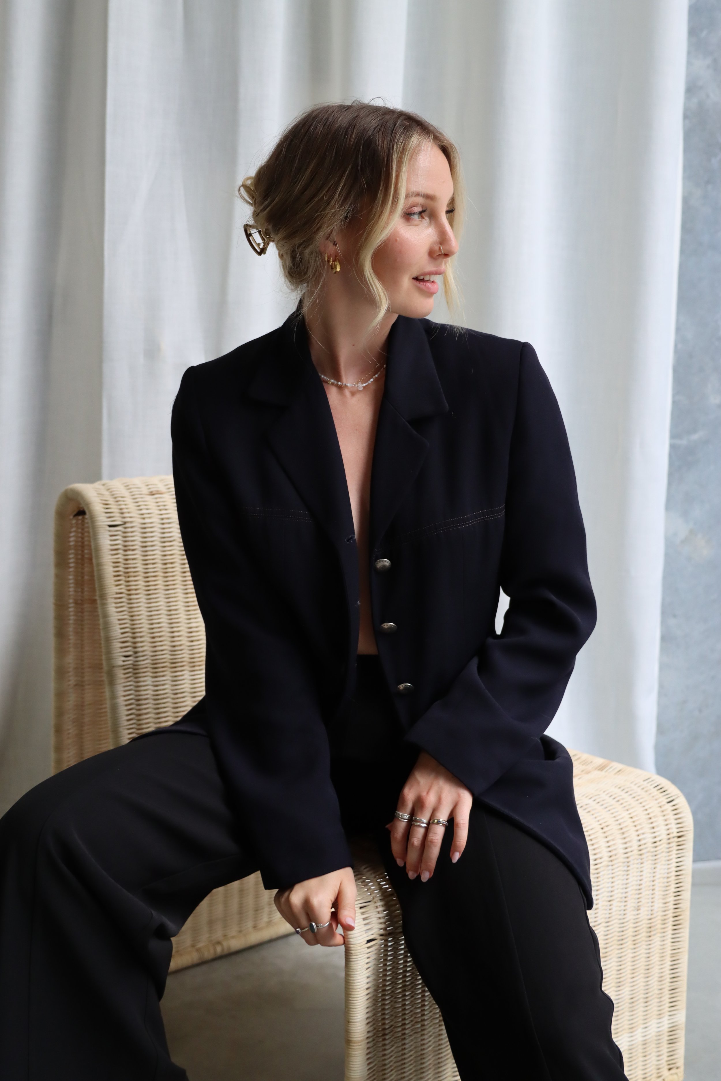 A woman with wavy blonde hair, wearing a black blazer and black pants, sits on a woven chair in front of light-colored curtains, looking to her left.