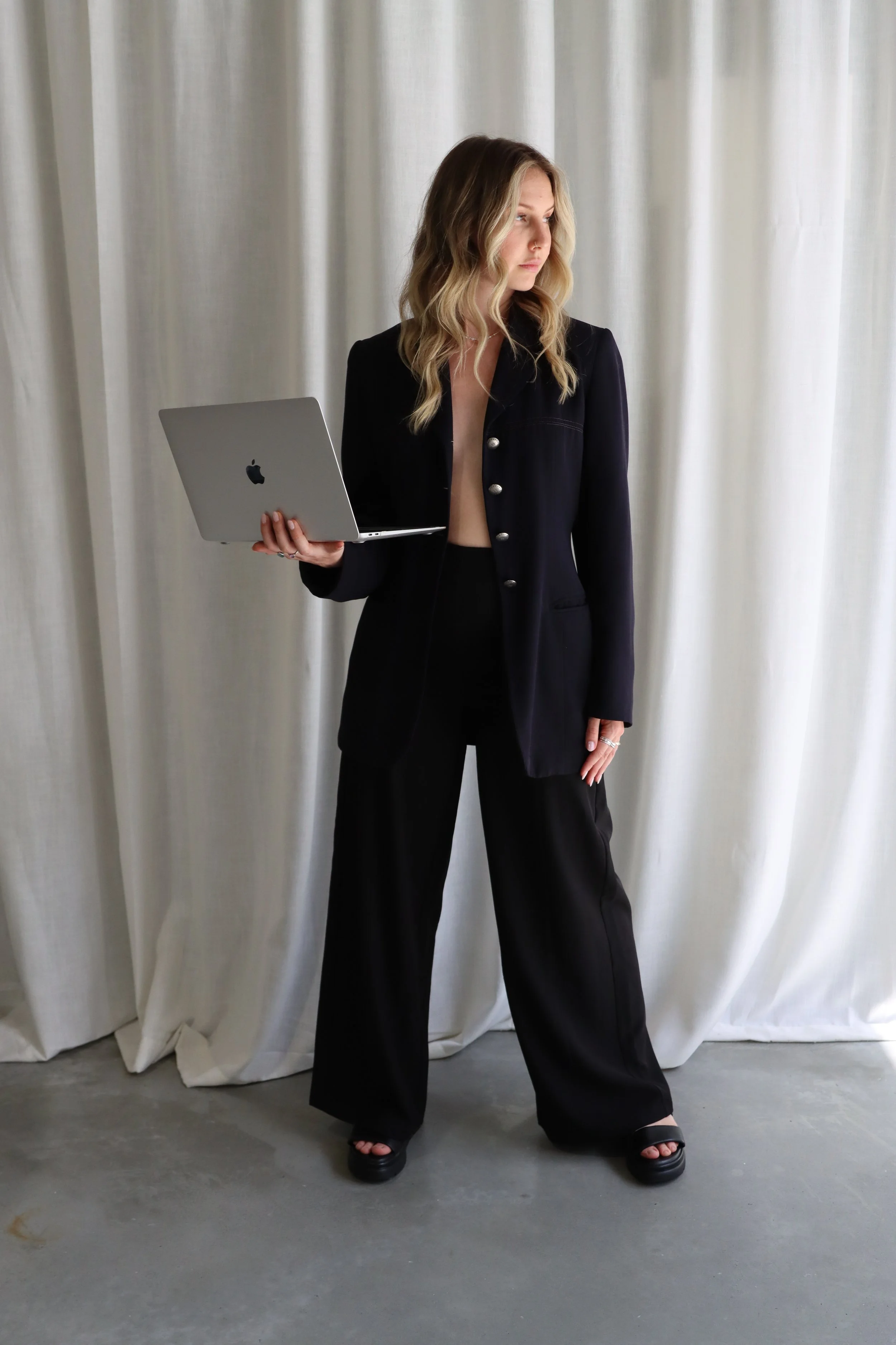 Woman with blonde wavy hair standing indoors, holding a silver MacBook, wearing a black blazer with silver buttons and black wide-leg pants, in front of light-colored curtains.