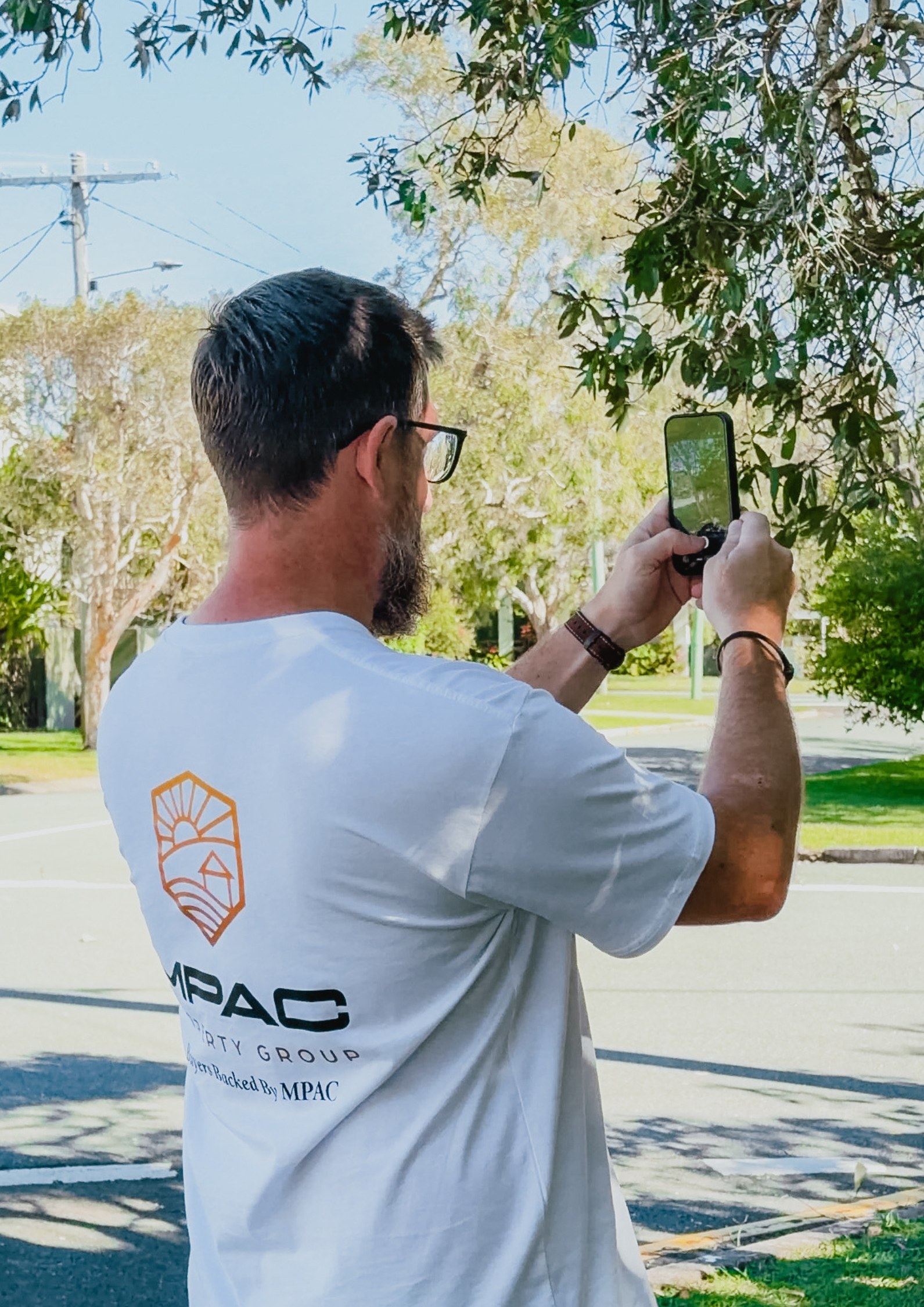 A man with dark hair, glasses, and a beard is standing outdoors on a sunny day, taking a photo or video with a smartphone. He is wearing a white MPAC Property Group t-shirt