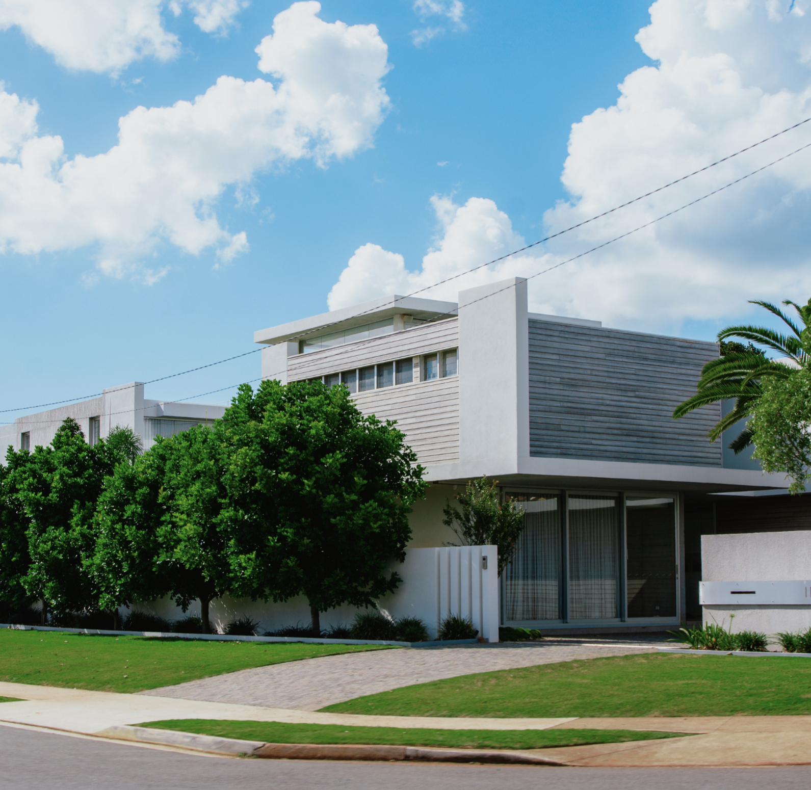 A modern white residential building with large glass doors and windows, surrounded by green trees, under a bright blue sky with scattered white clouds.