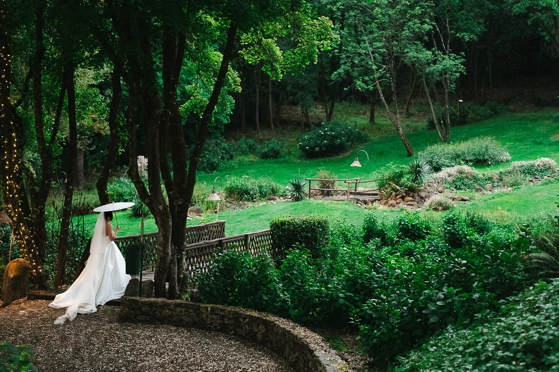 Una donna in abito da sposa con ombrello bianco cammina lungo un sentiero nel giardino verde, circondata da alberi e arbusti, in una scena serale o piovosa.
