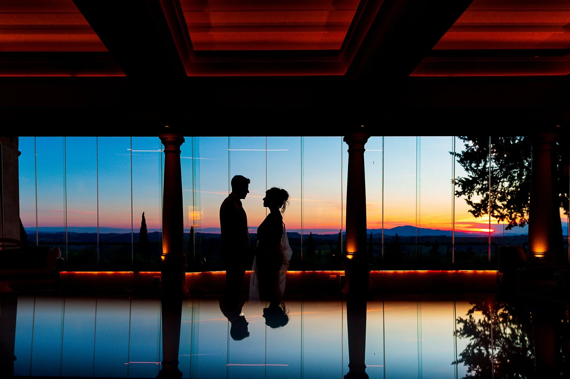 Silhouette di una coppia innamorata che si tiene per mano in una stanza con grandi finestre, al tramonto, con vista sul paesaggio collinare.